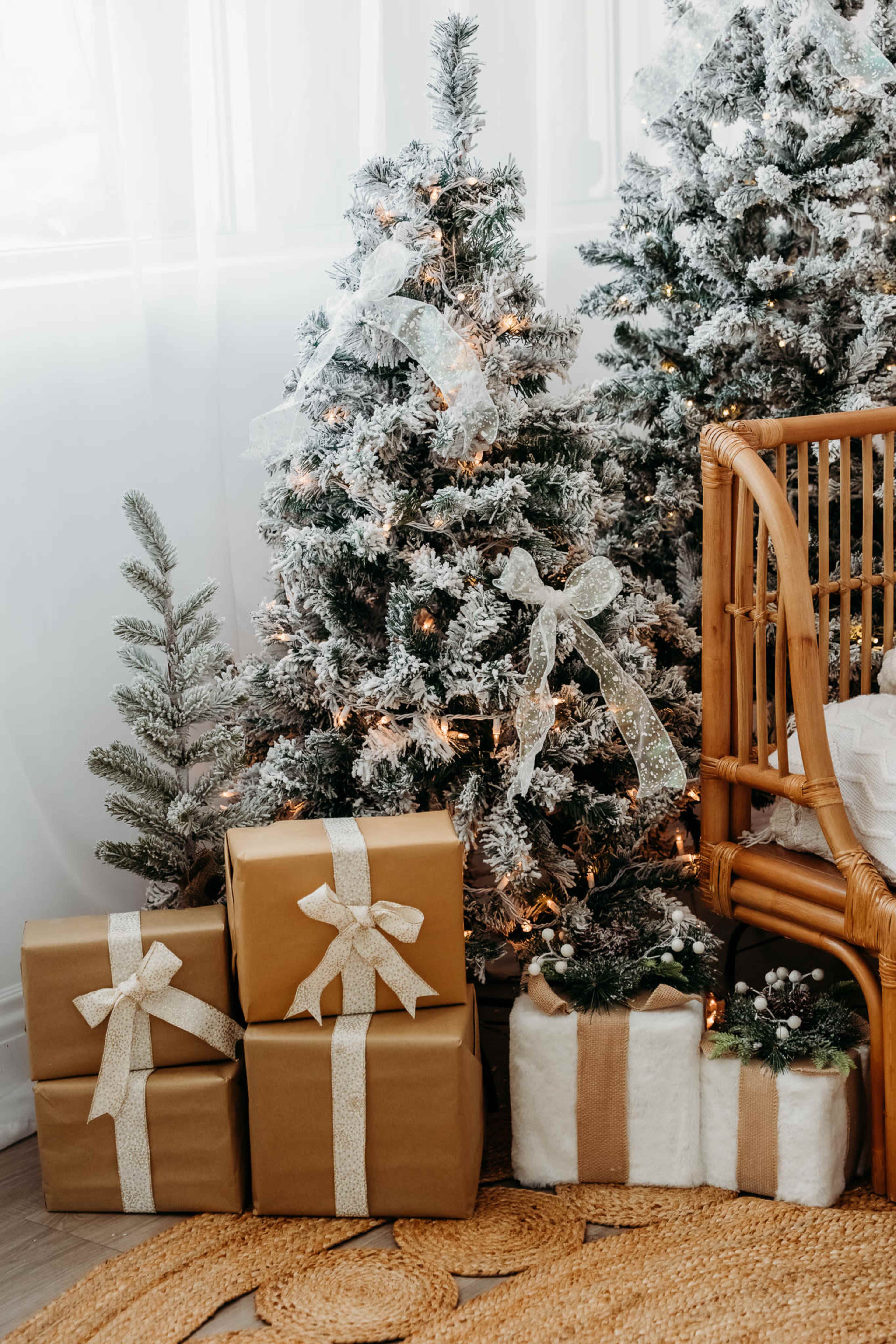 The scene features two frosted Christmas trees adorned with bows and lights, accompanied by neatly wrapped presents in brown and white, placed beside a wicker chair and a jute rug.