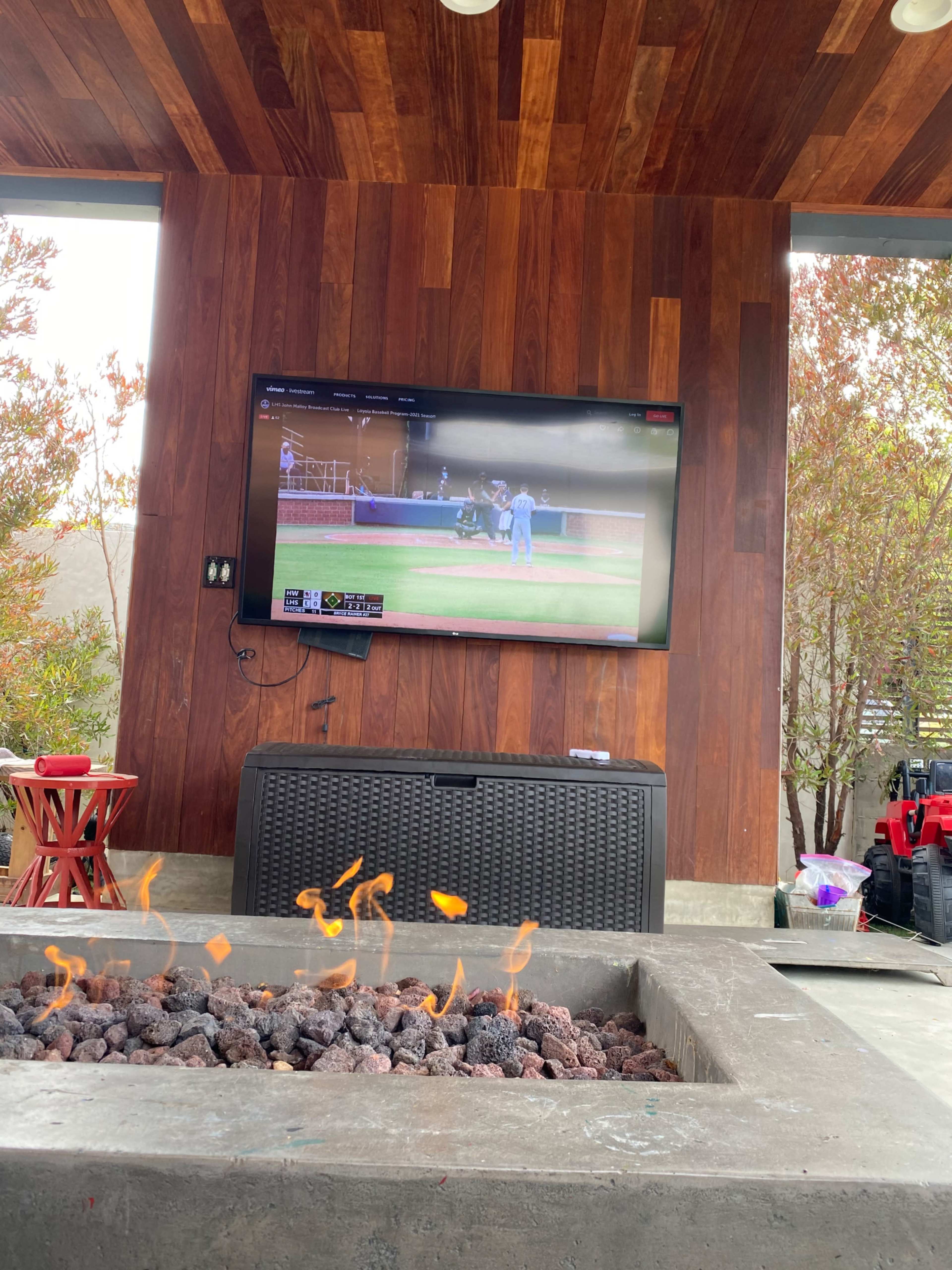 A fire pit with visible flames is in the foreground, while a television mounted on a wooden wall shows a baseball game in an outdoor seating area.