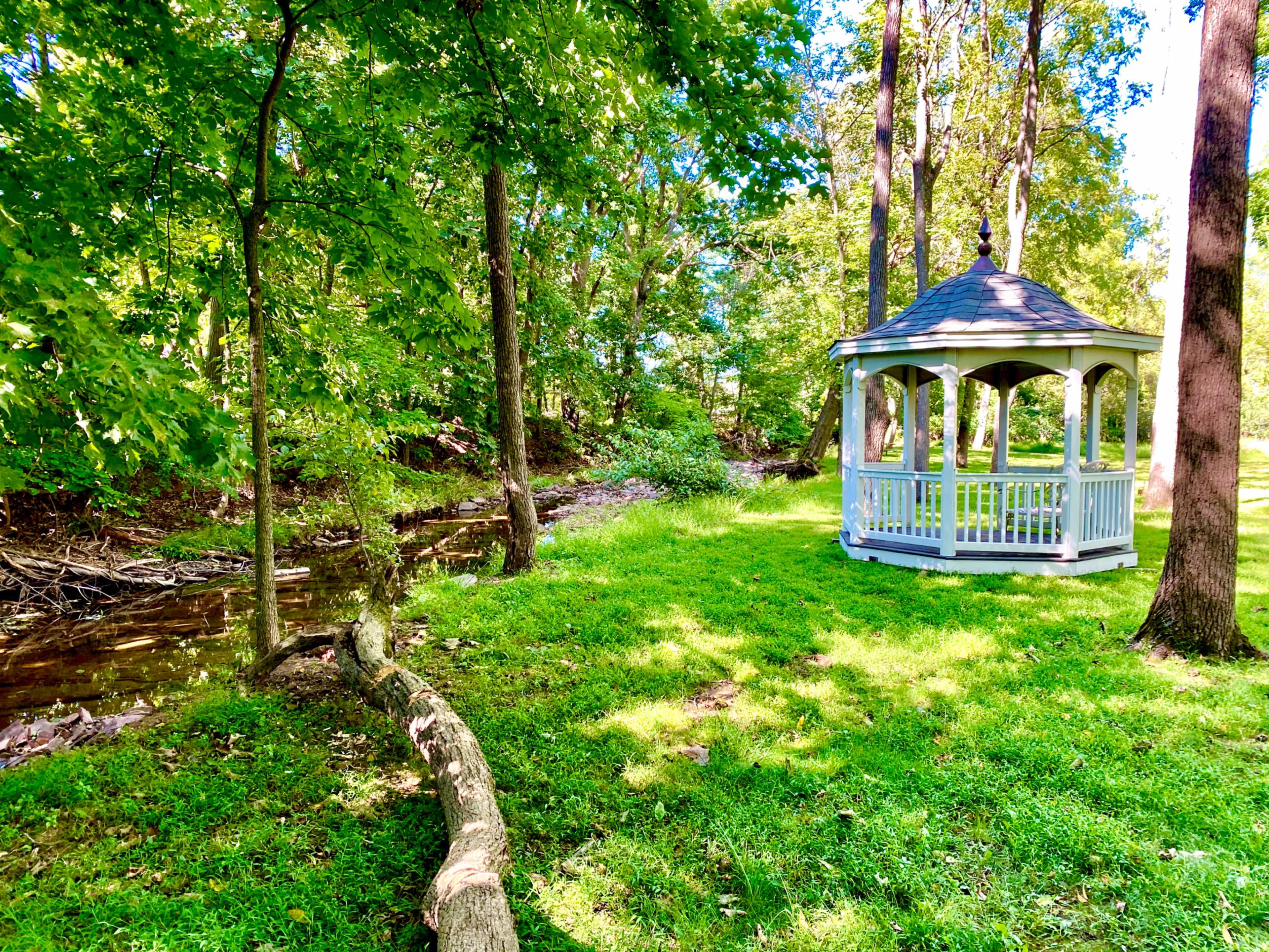 A white gazebo stands on a grassy area beside a small creek, surrounded by tall trees.
