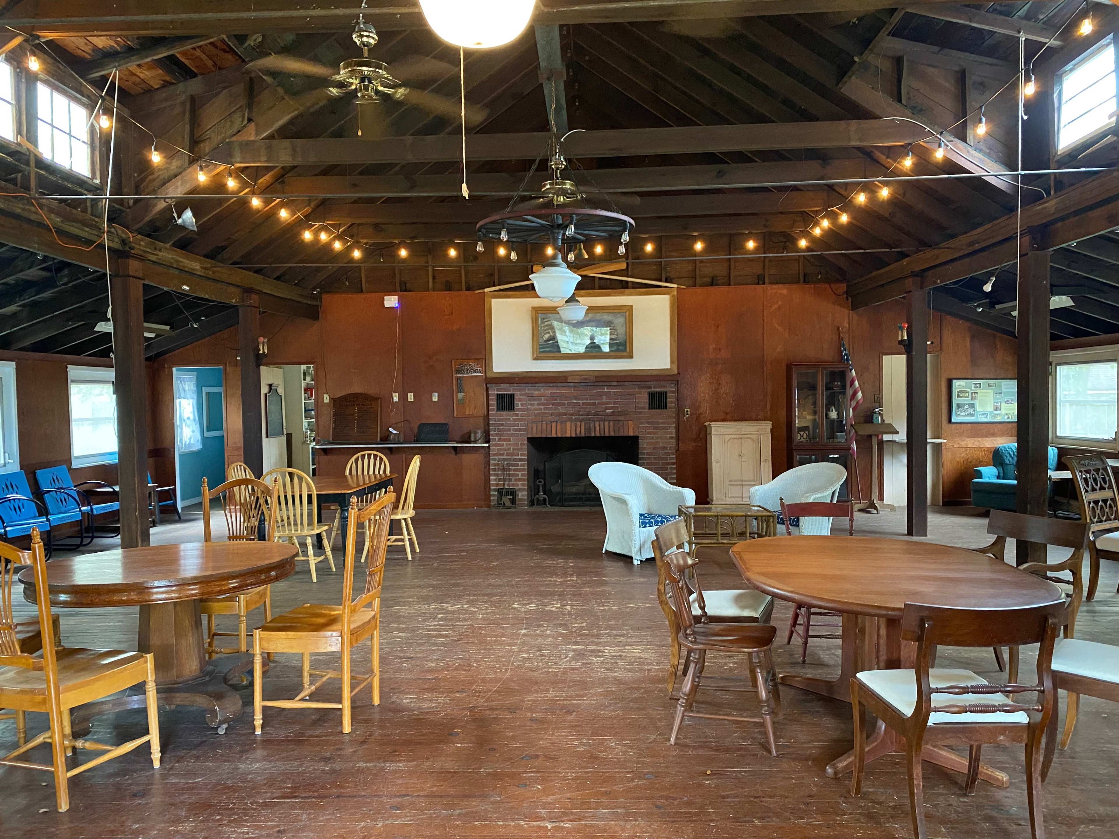 The image shows a spacious, wooden interior of a lodge with tables and chairs arranged in the open area, a fireplace in the back, and string lights hanging from the ceiling.