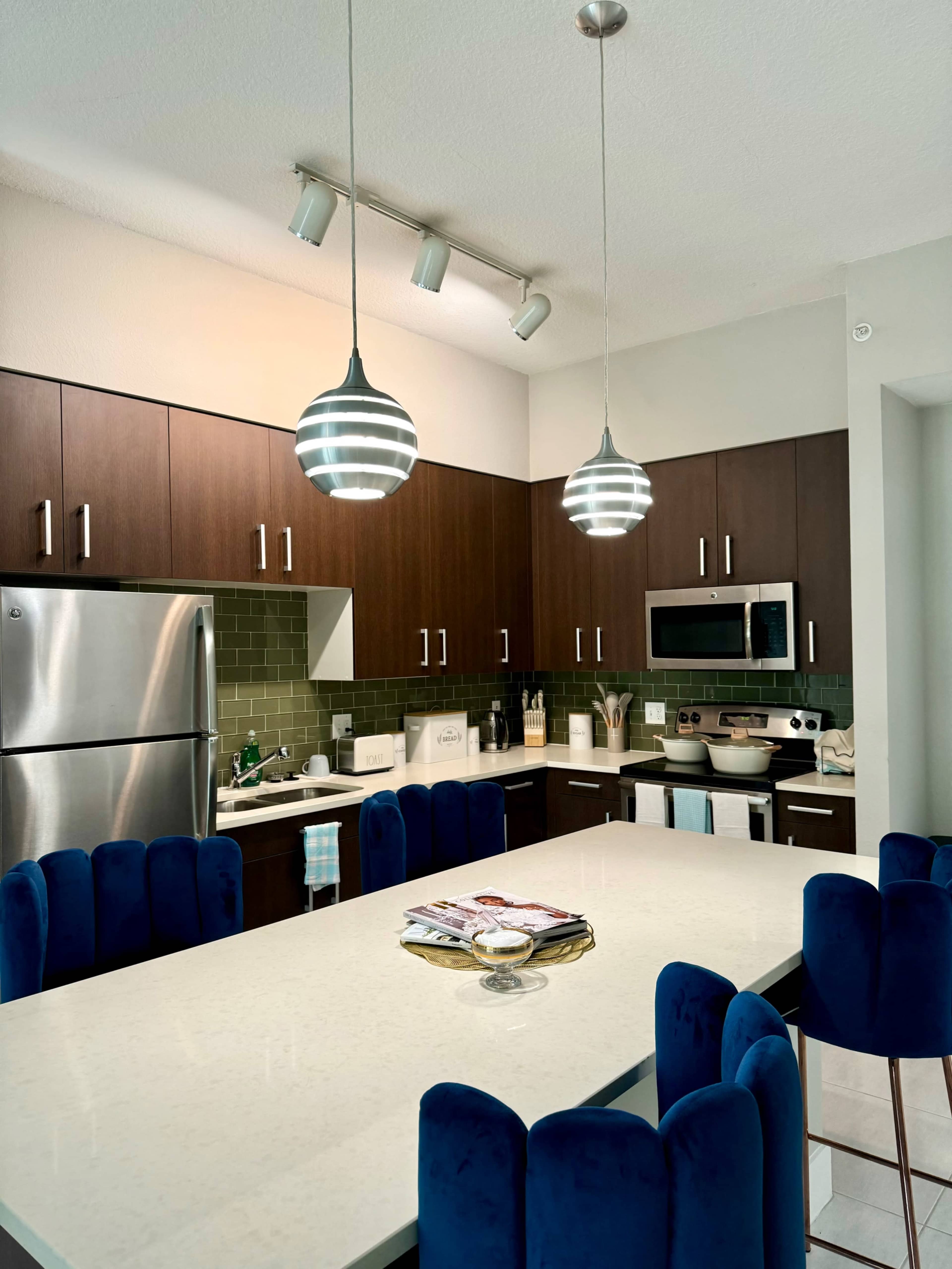 The kitchen features dark wood cabinets, stainless steel appliances, and a central island with blue upholstered chairs and modern pendant lighting.
