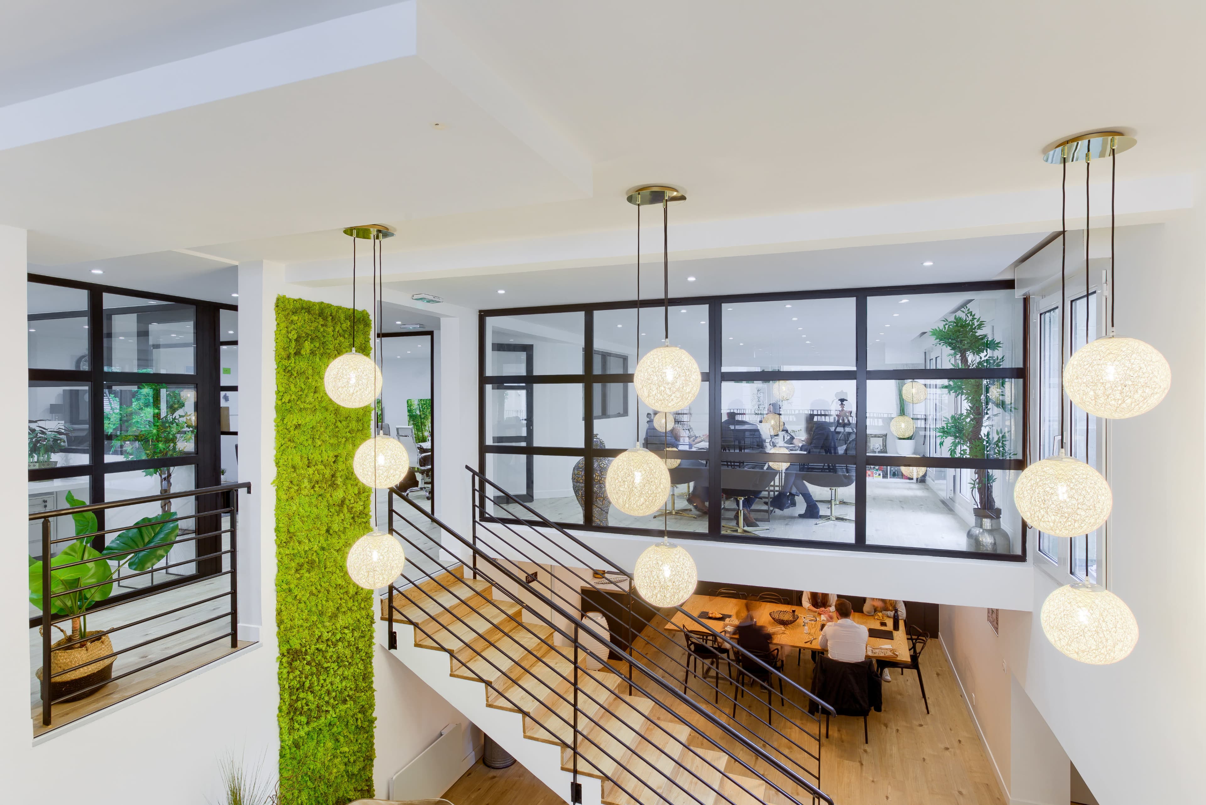 A modern interior features a staircase with hanging spherical lights, surrounded by greenery and large windows overlooking a dining area.