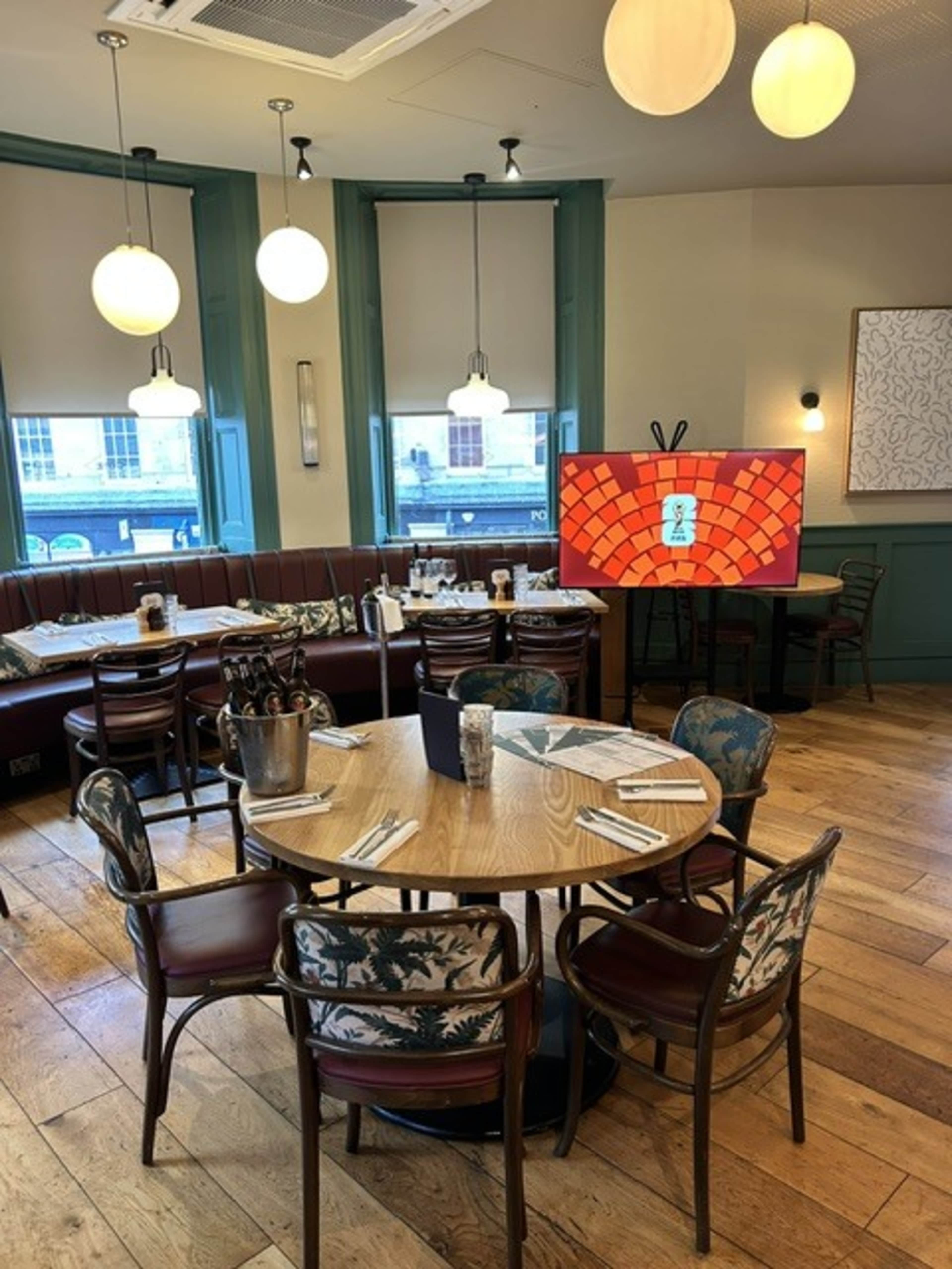 The image shows an inviting restaurant interior with round wooden tables, upholstered chairs, and a large screen displaying a colorful graphic.