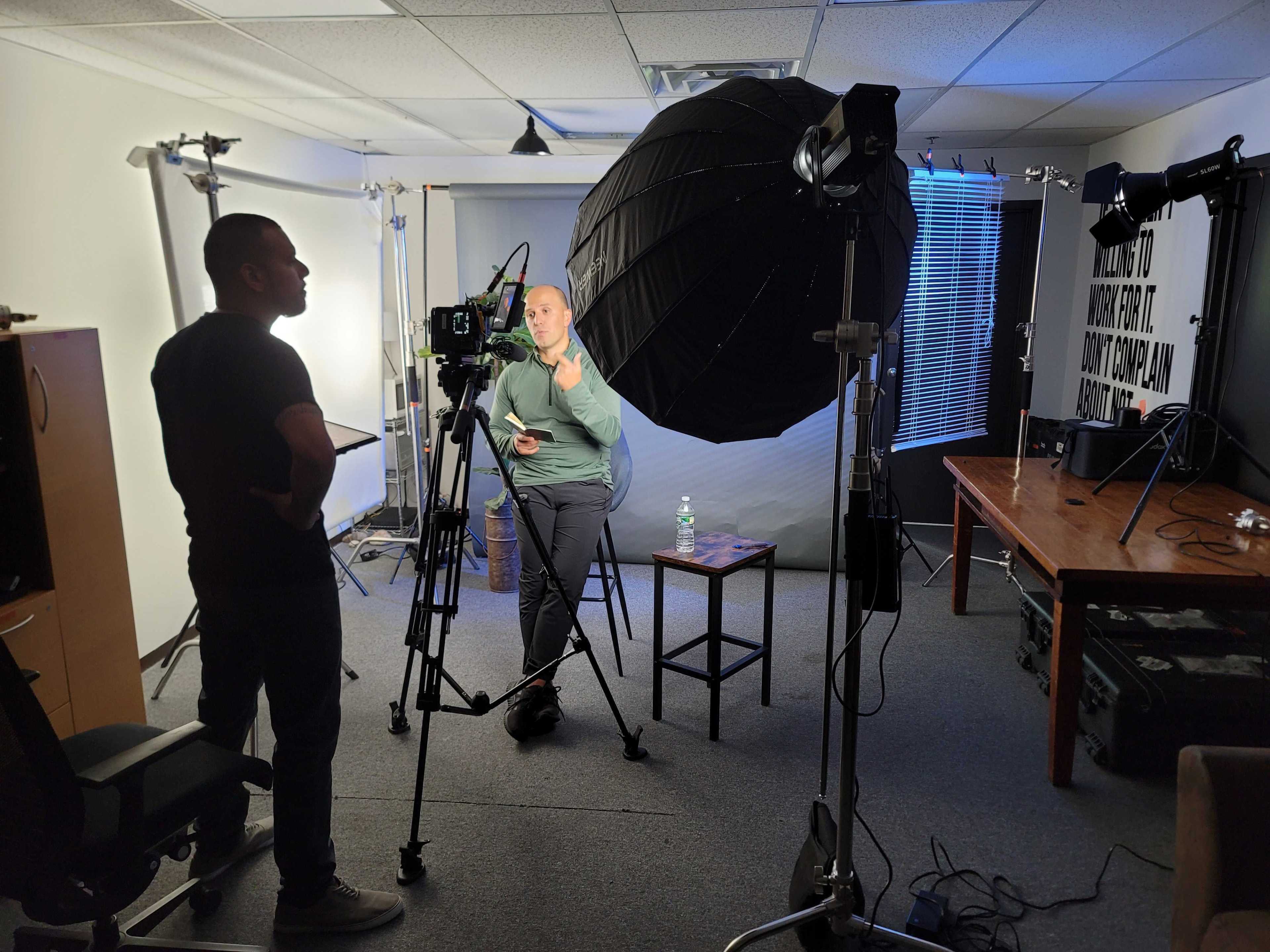 A person stands in front of a camera setup with a large softbox light in a well-equipped studio.