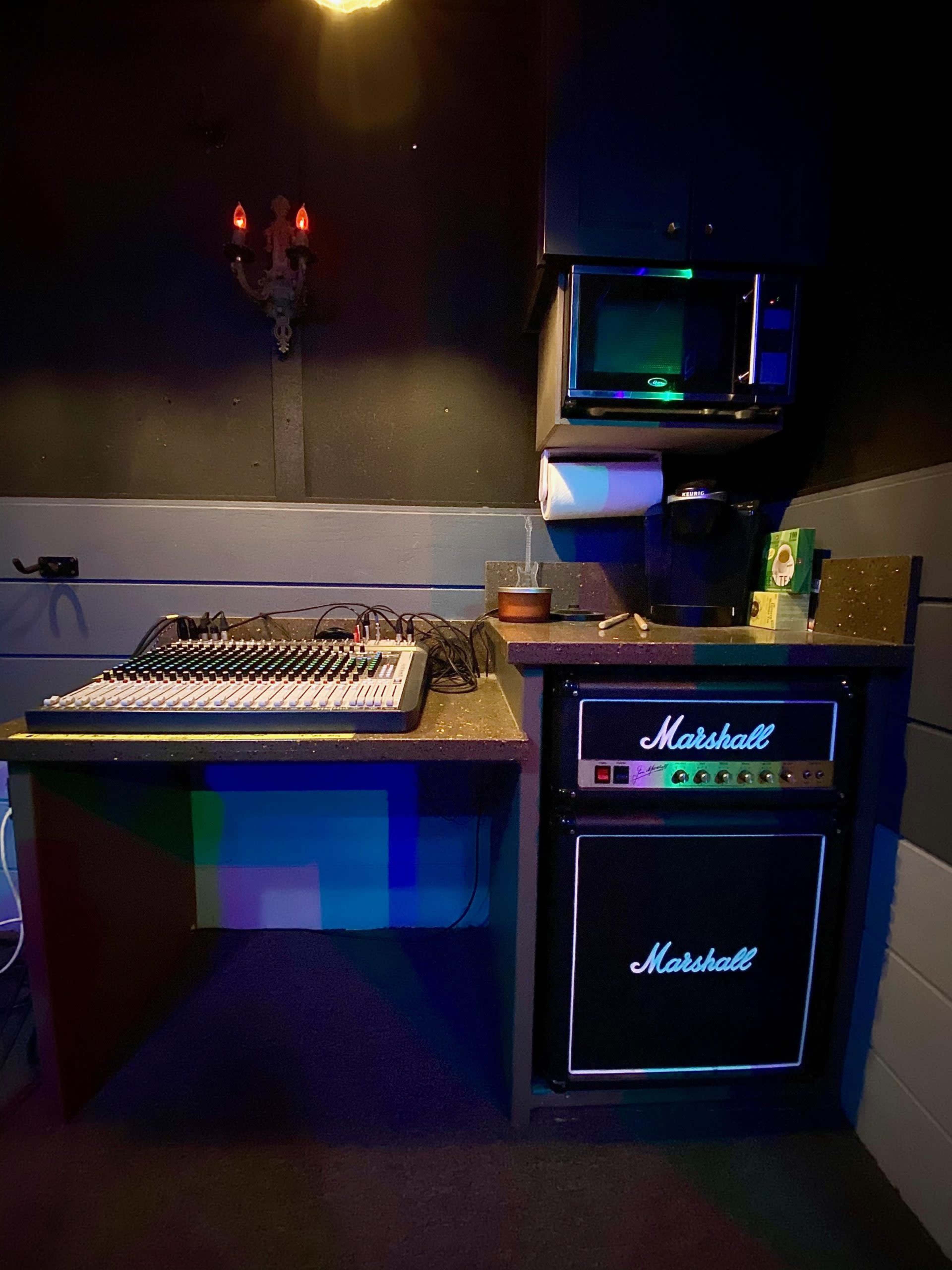 A sound mixing console placed on a table next to a Marshall amplifier and various kitchen appliances in a dimly lit room.