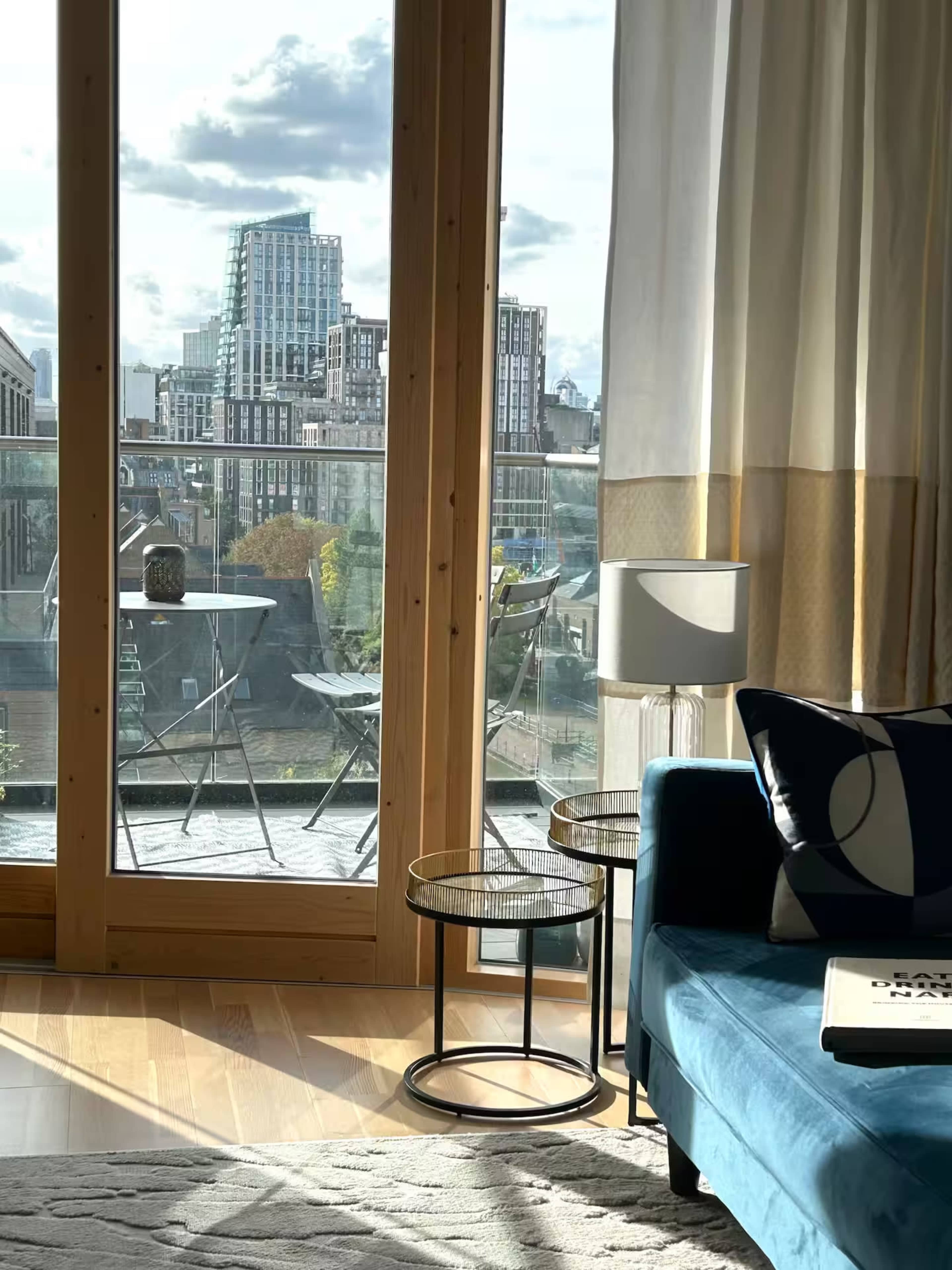 A sunlight-filled living room features a blue couch, two small tables, and large windows overlooking a cityscape.