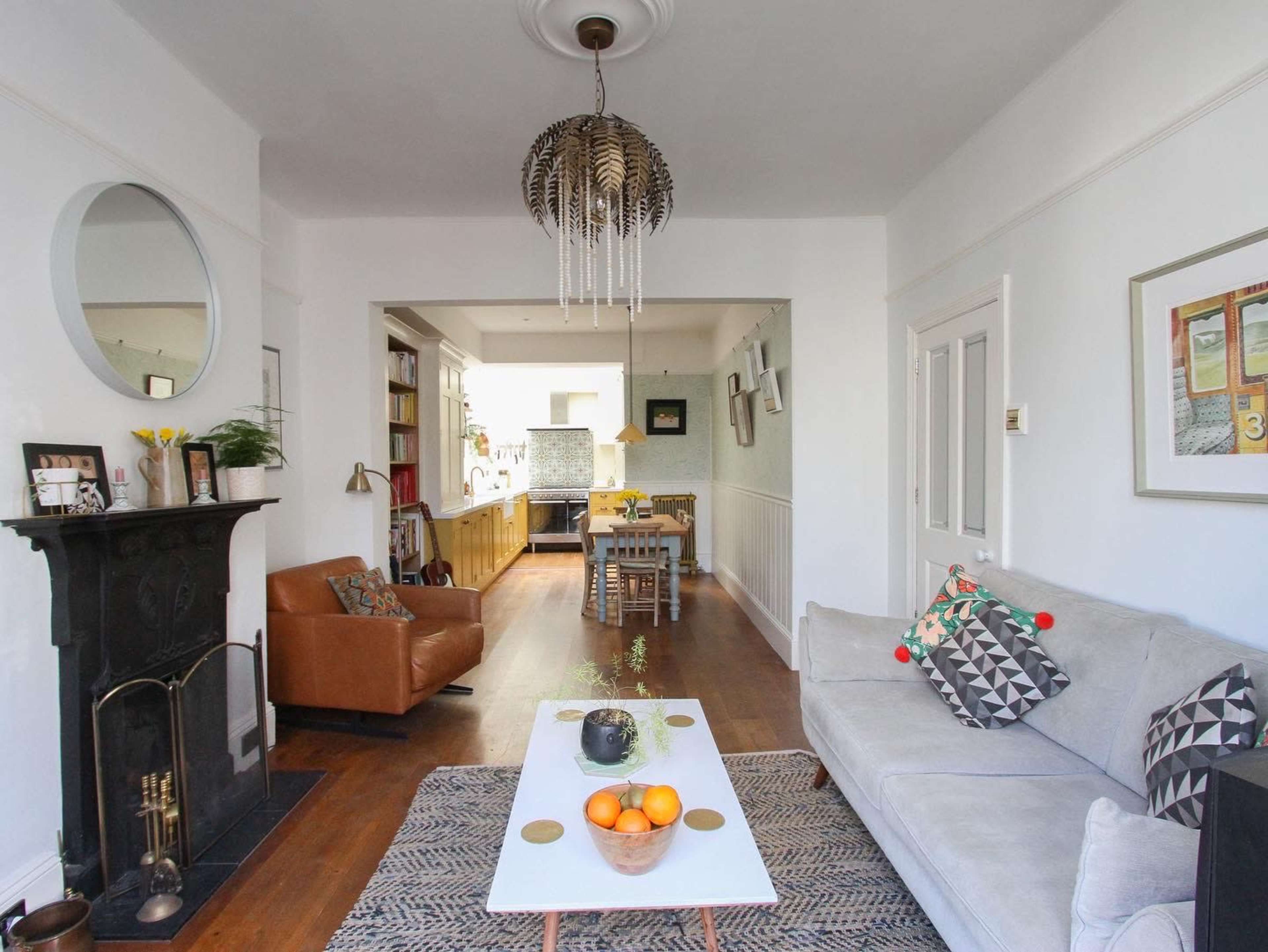 A living room features a sofa, coffee table with oranges, a fireplace, and an adjoining kitchen, all set against light-colored walls and wooden floors.
