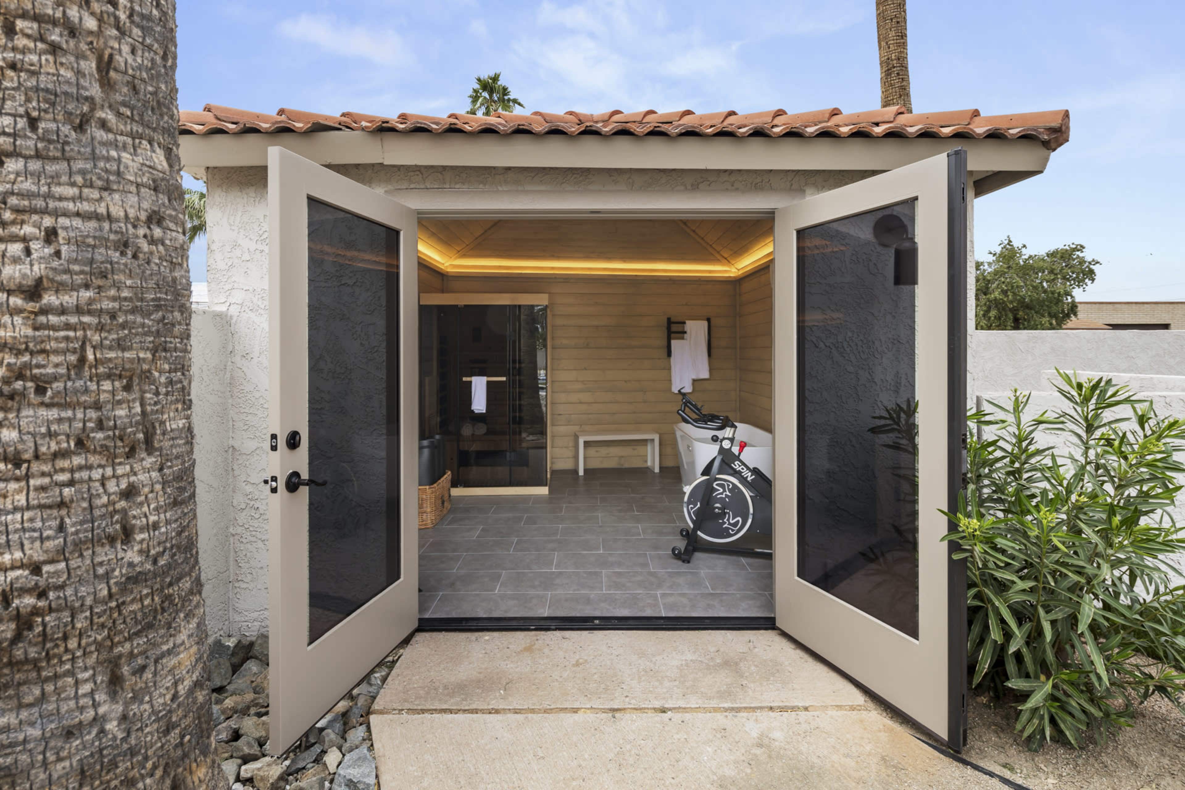 The image shows a modern outdoor gym with large glass doors opening to an interior featuring exercise equipment and warm lighting.