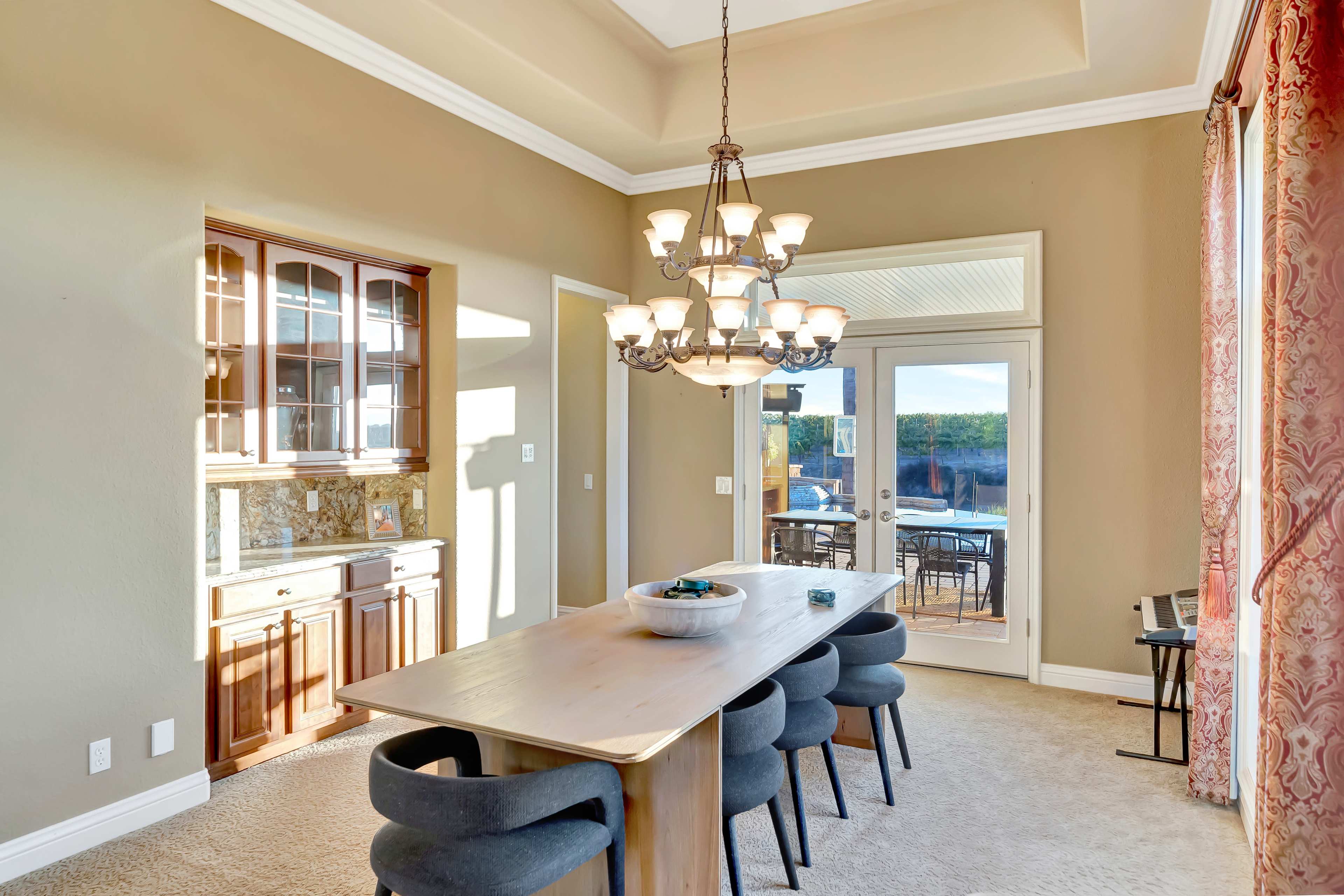 A spacious dining area features a long wooden table surrounded by upholstered chairs, with a chandelier above and a view of an outdoor patio through sliding glass doors.