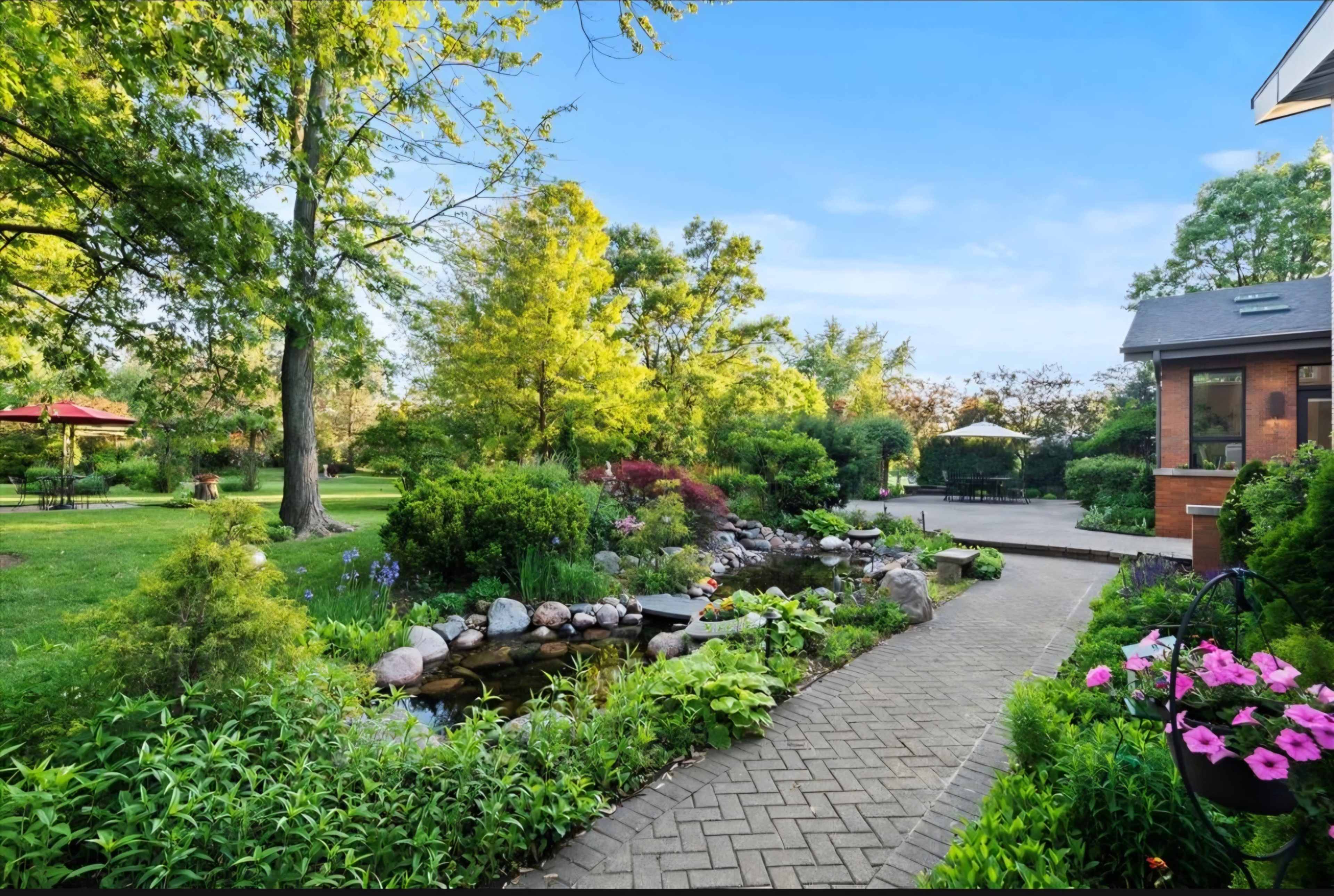 The image shows a landscaped garden with a stone pathway leading to a pond, surrounded by various plants and trees.