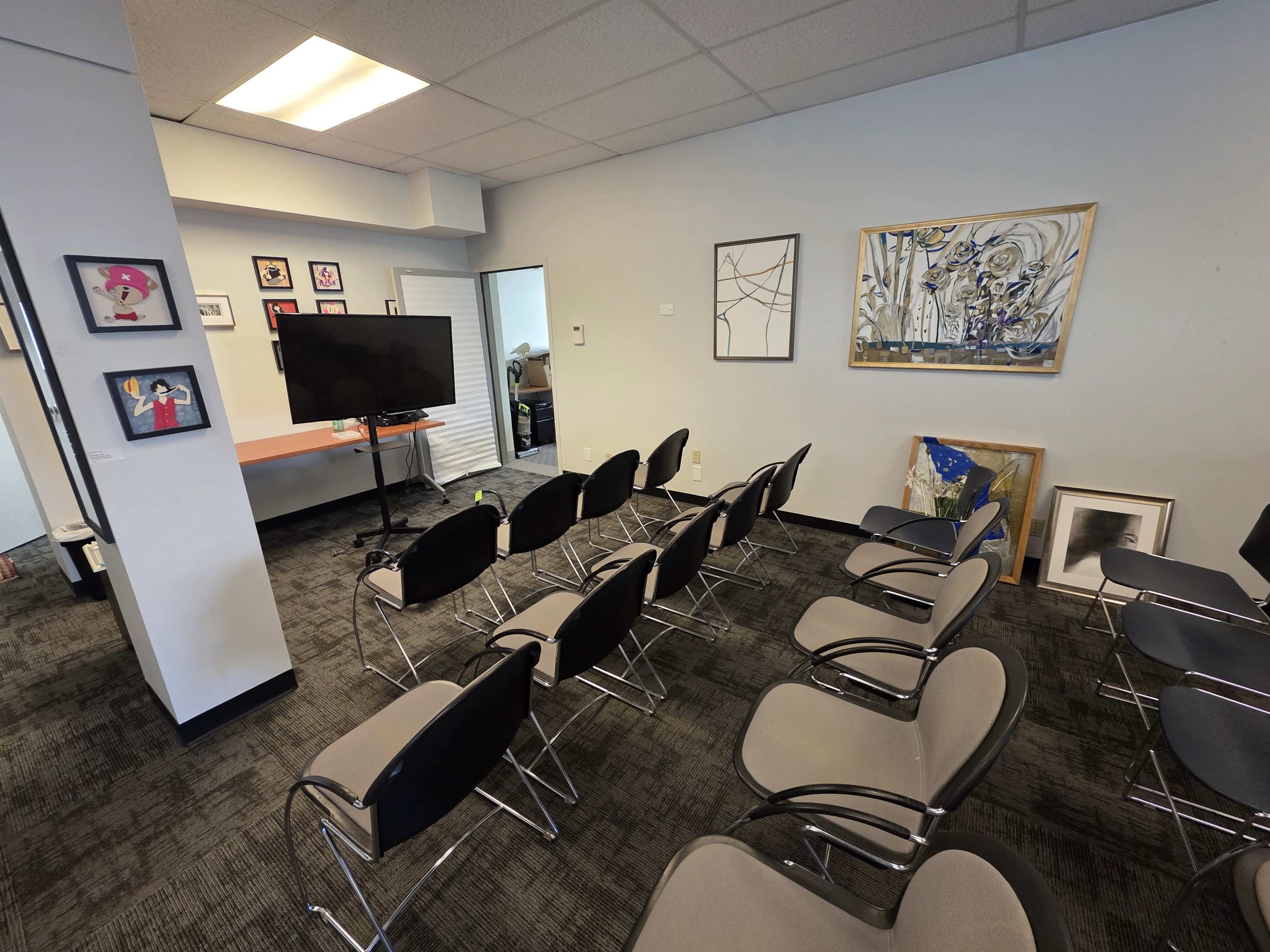 The image shows a modest conference room with rows of chairs facing a television screen mounted on a wall, accented by framed artworks and a small table.