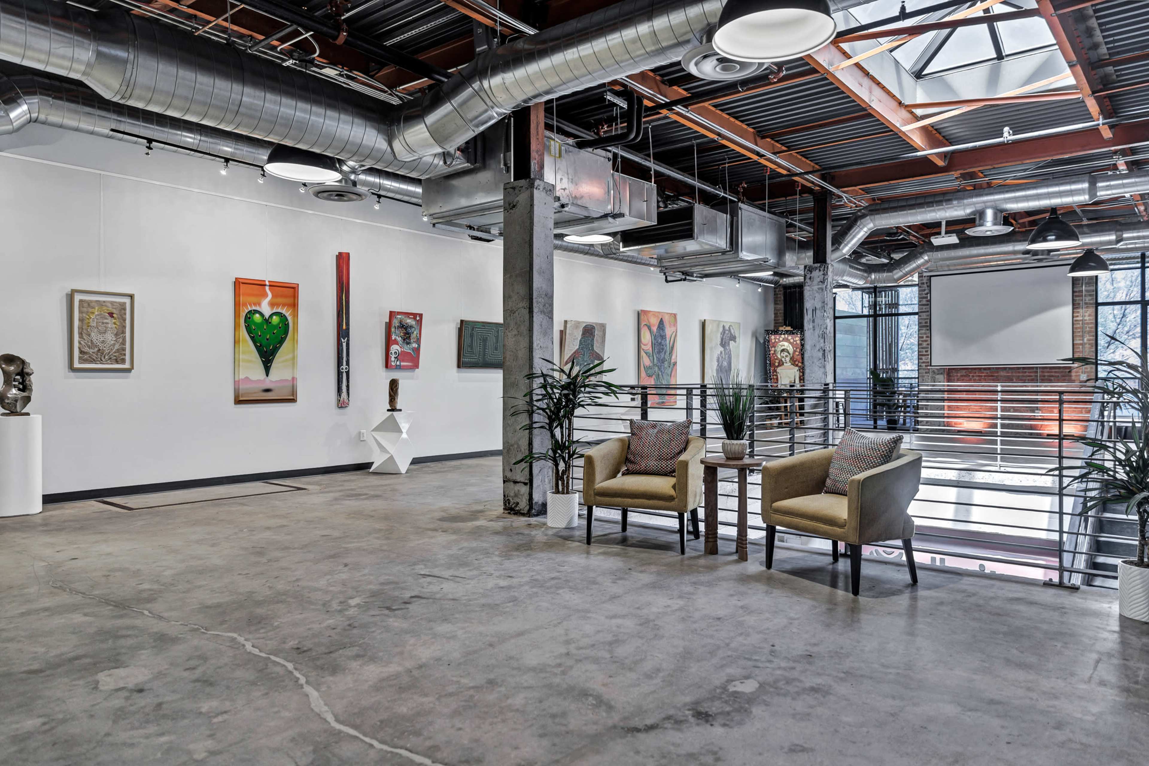 The image shows an art gallery with a spacious interior featuring various artworks on the walls, modern seating, and a large window allowing natural light to enter.