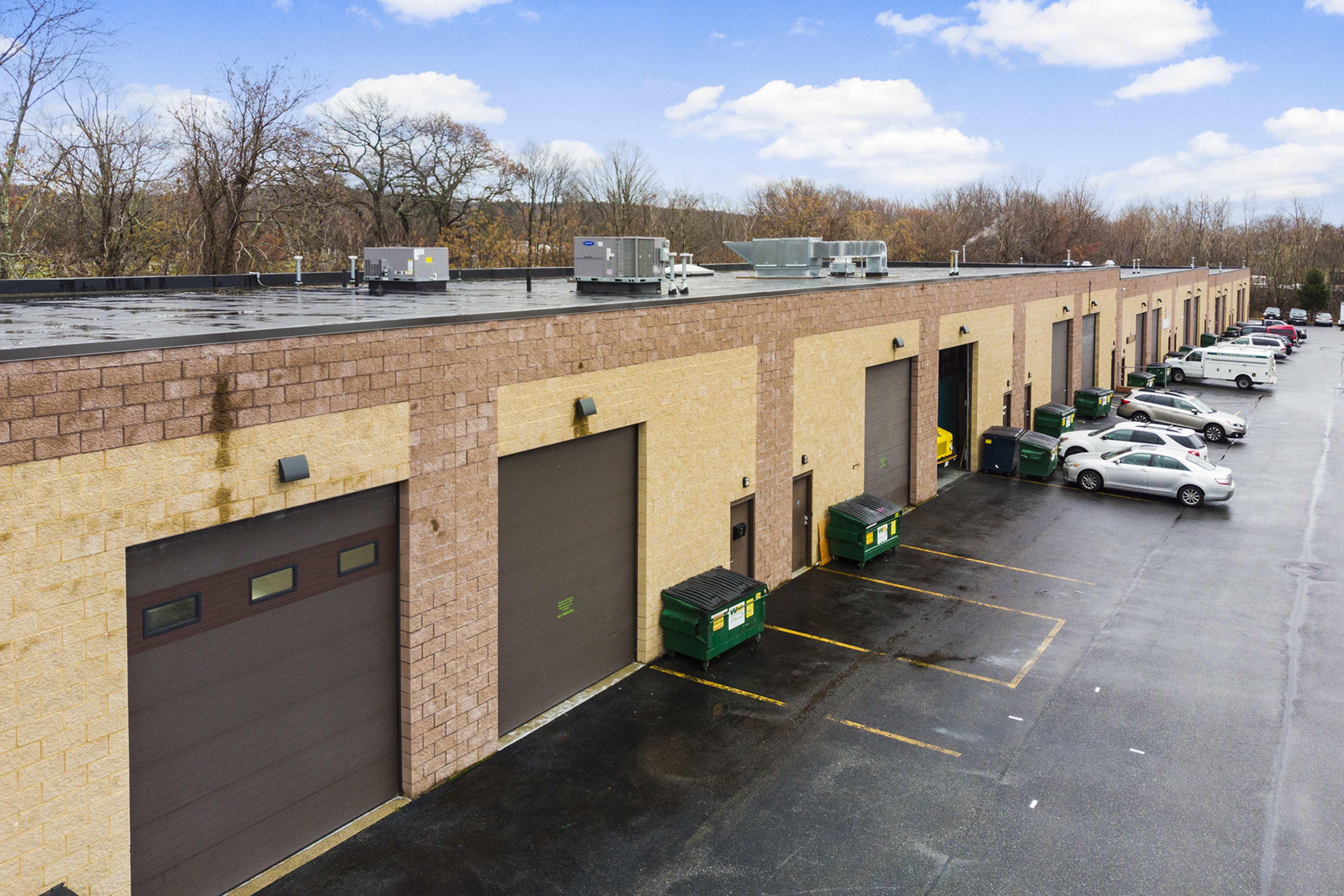 A row of industrial garage doors on a commercial building, with parked cars and green dumpsters lined along the side.