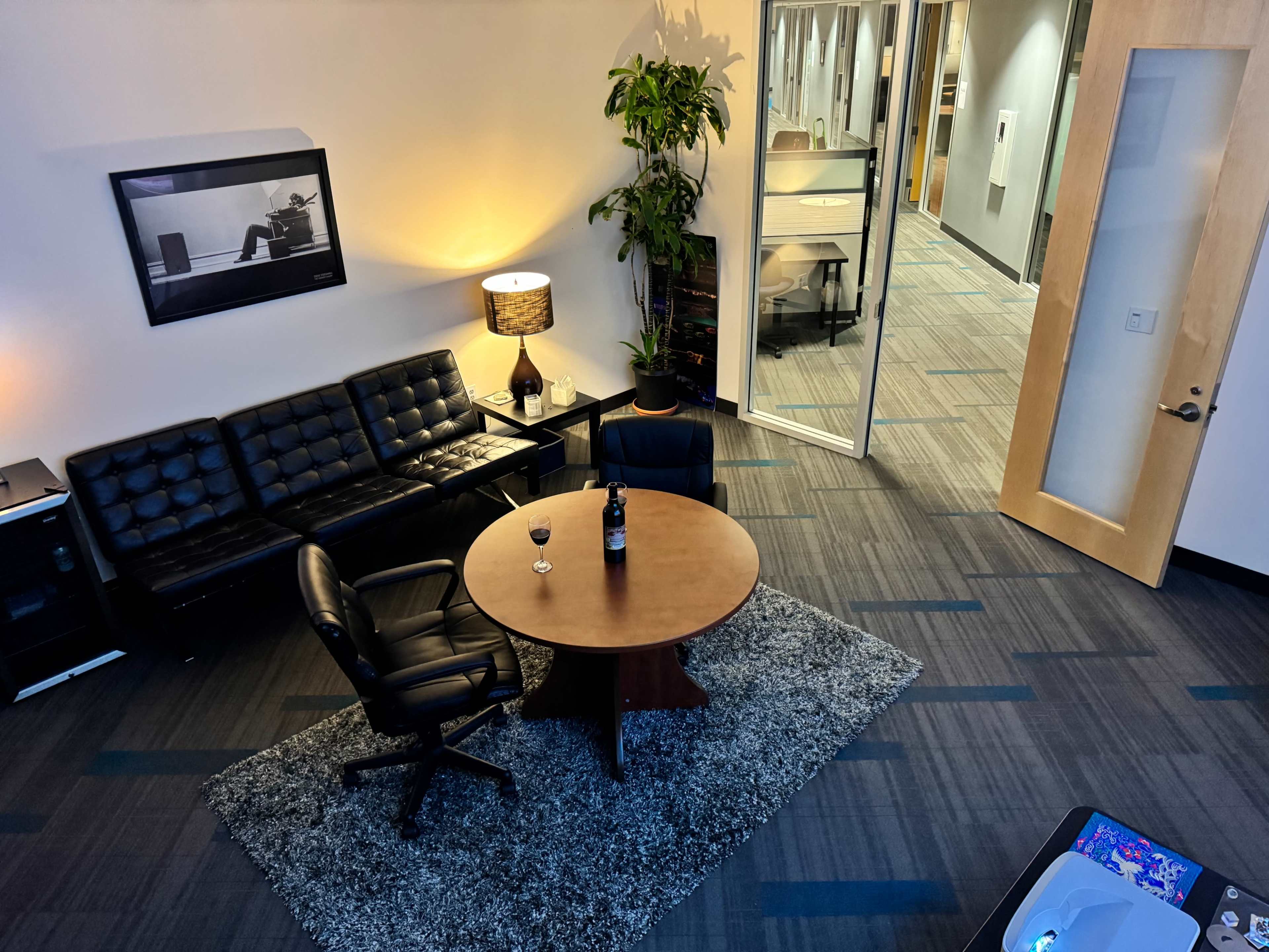 A modern office lounge area featuring a round wooden table, leather chairs, a sofa, and a glass of wine on the table, with a plant in the corner and an open doorway leading to another workspace.