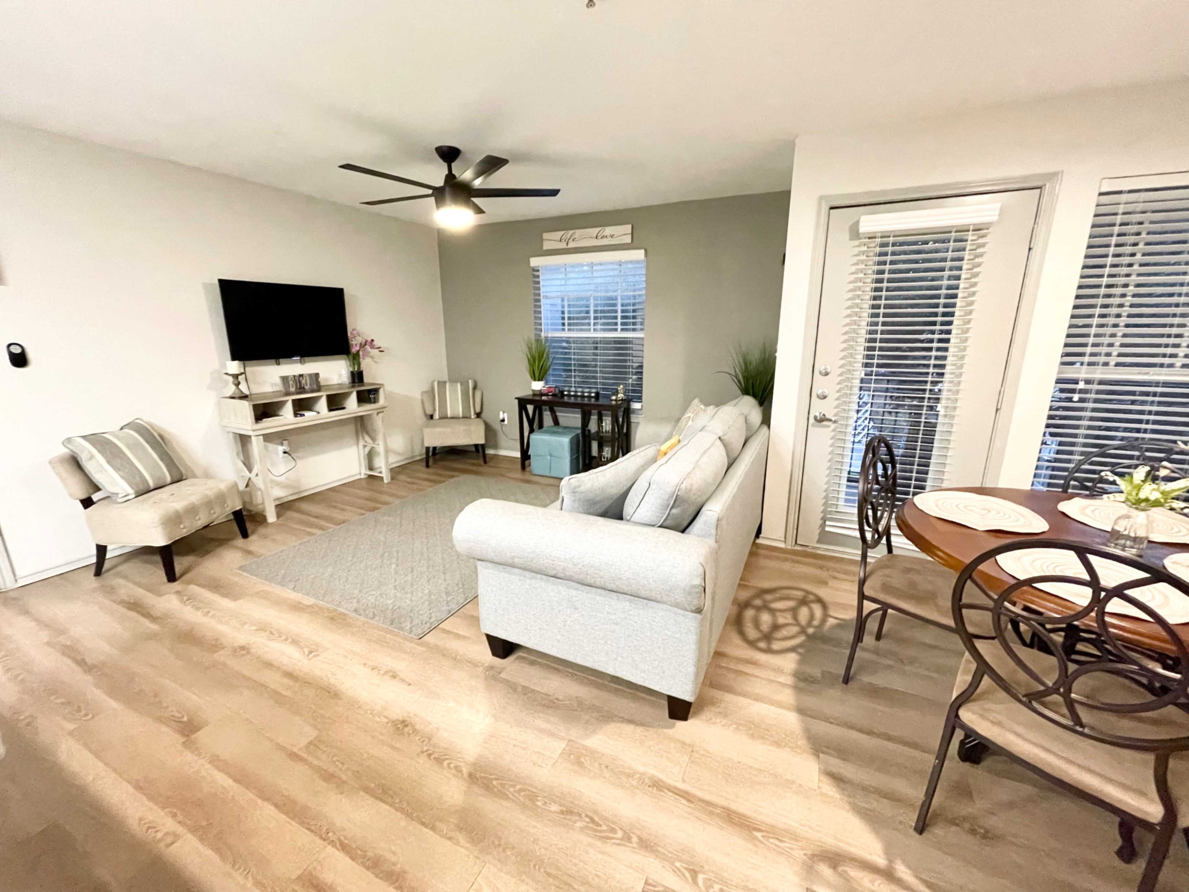 A living room with a light-colored sofa, a small dining table by the window, and a television mounted on the wall.