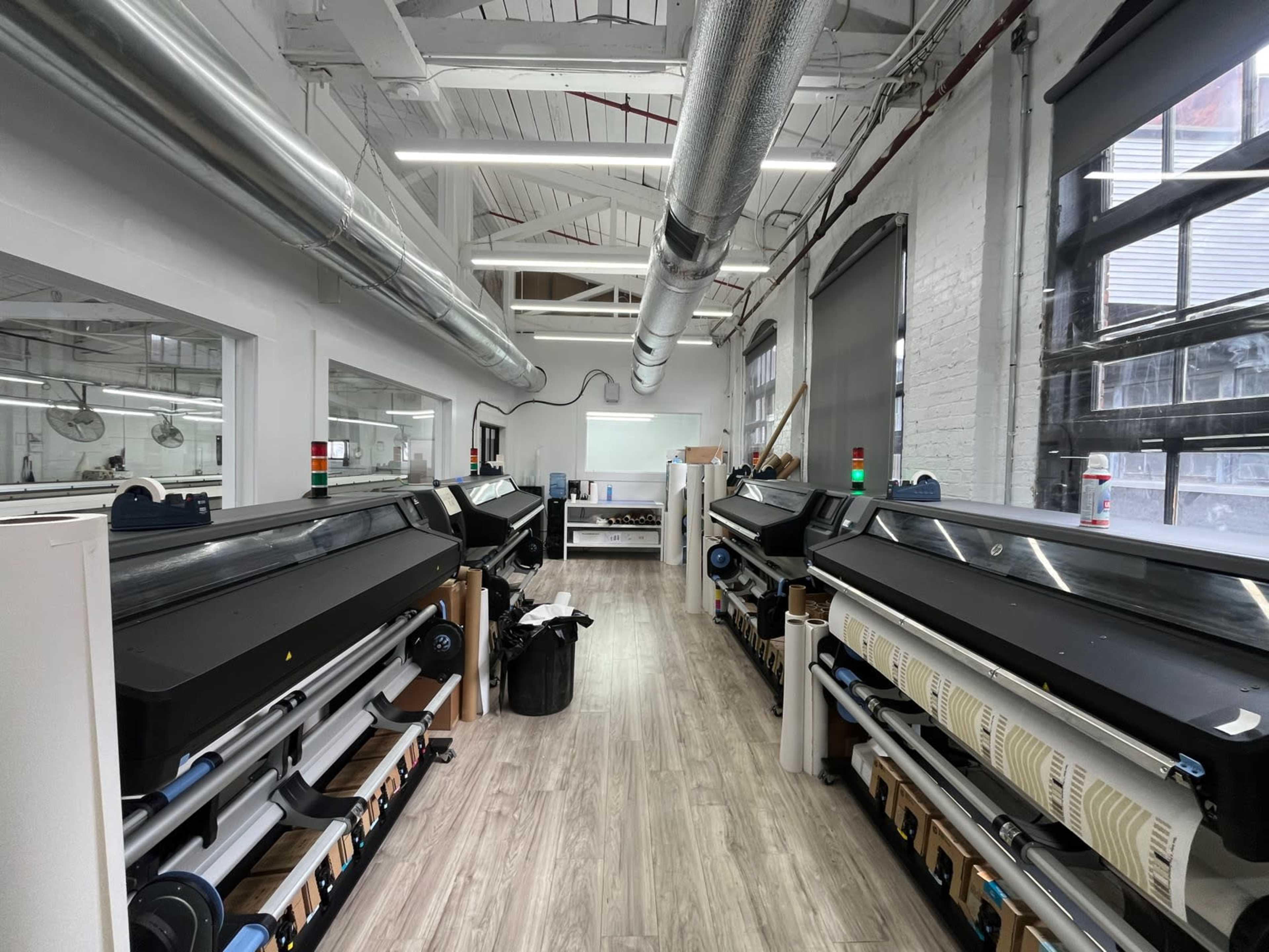 The image shows a large printing workspace with several printers aligned along the walls, surrounded by windows and industrial ventilation pipes.