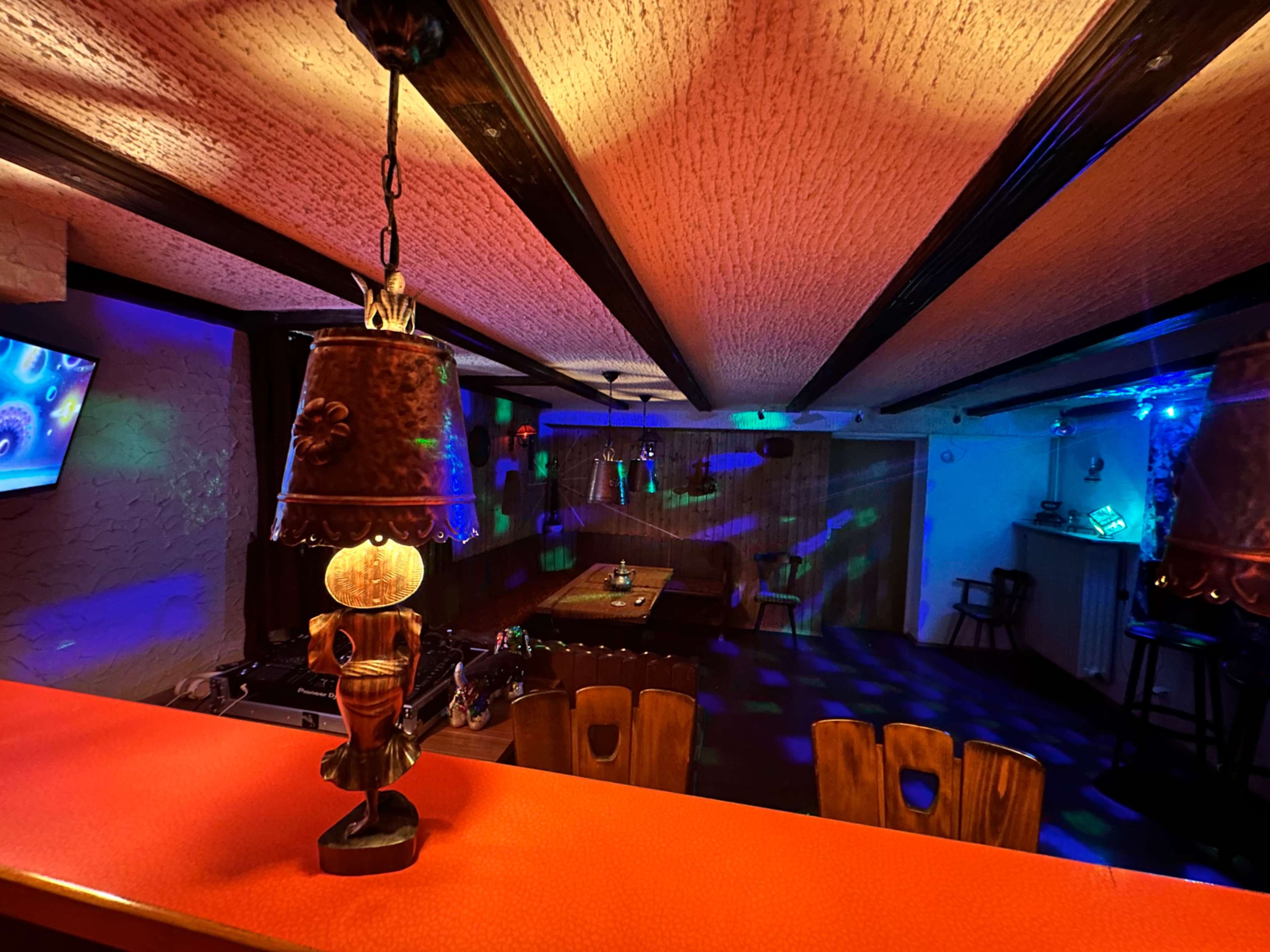 A dimly lit bar area with colorful lights reflecting on the walls and a pool table in the background.