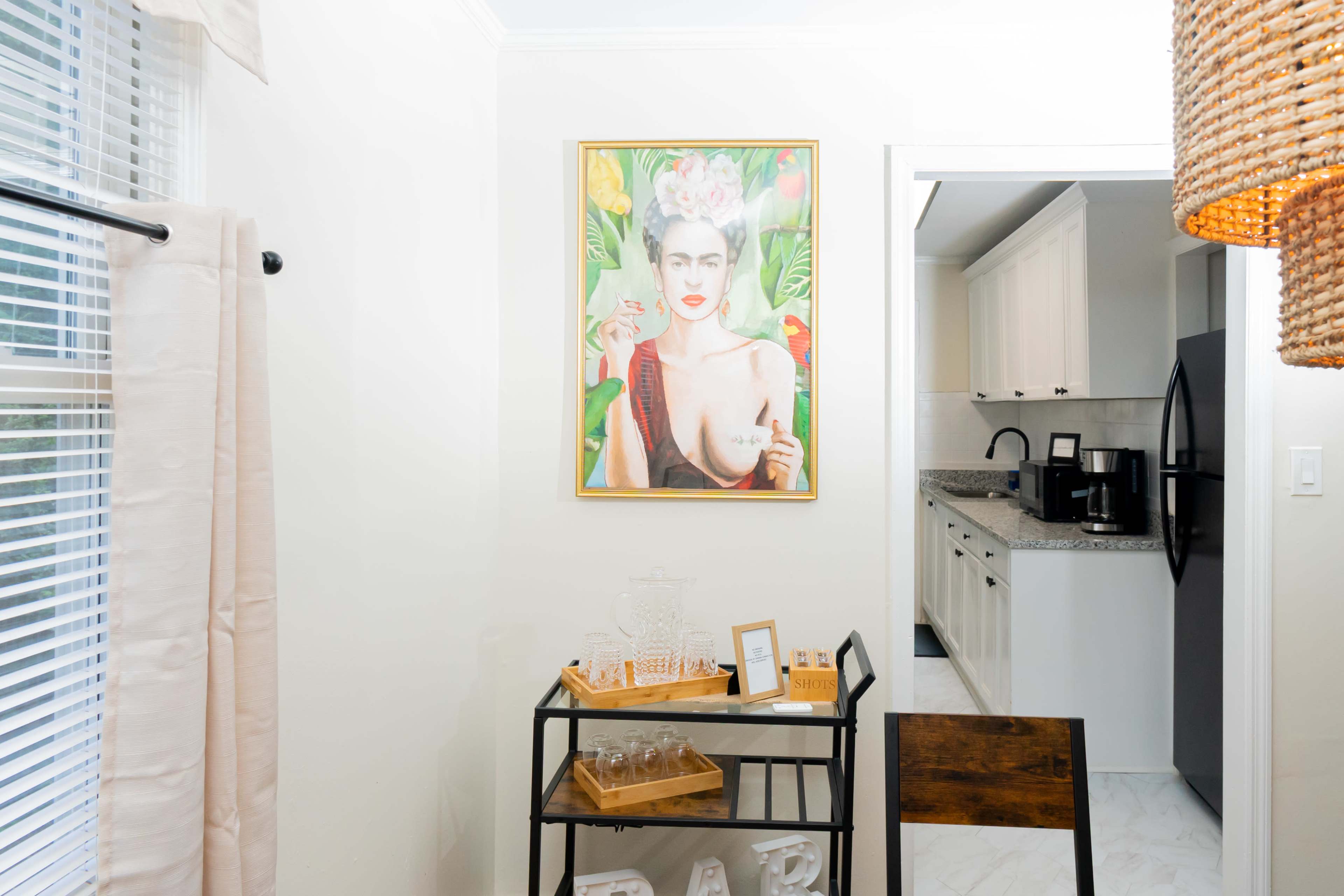 A framed portrait of a woman with flowers in her hair hangs on a light wall opposite a kitchen area with black appliances and white cabinets.