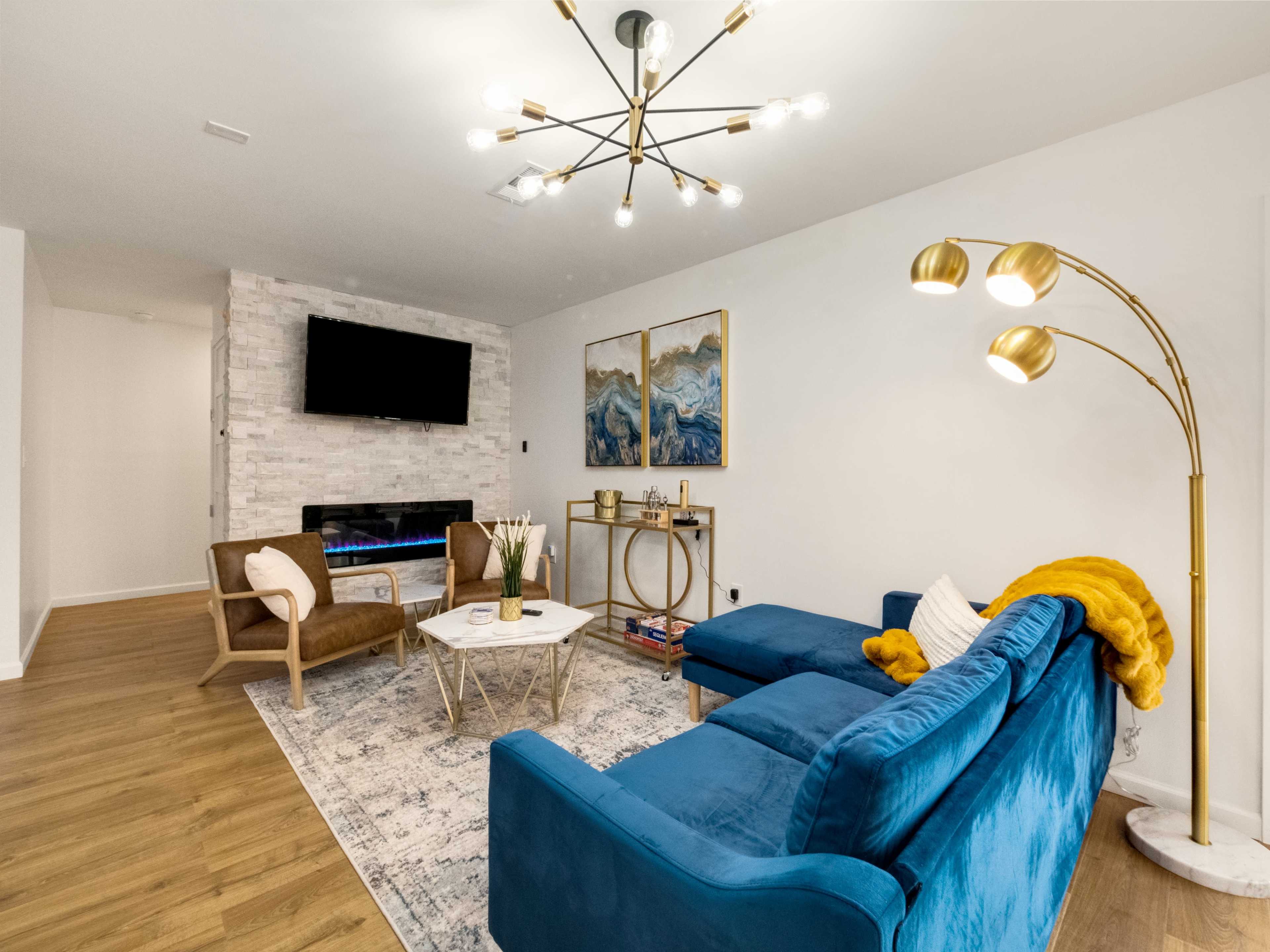 The living room features a blue sectional sofa, a gold floor lamp, a wall-mounted television, and a modern coffee table on a patterned area rug.