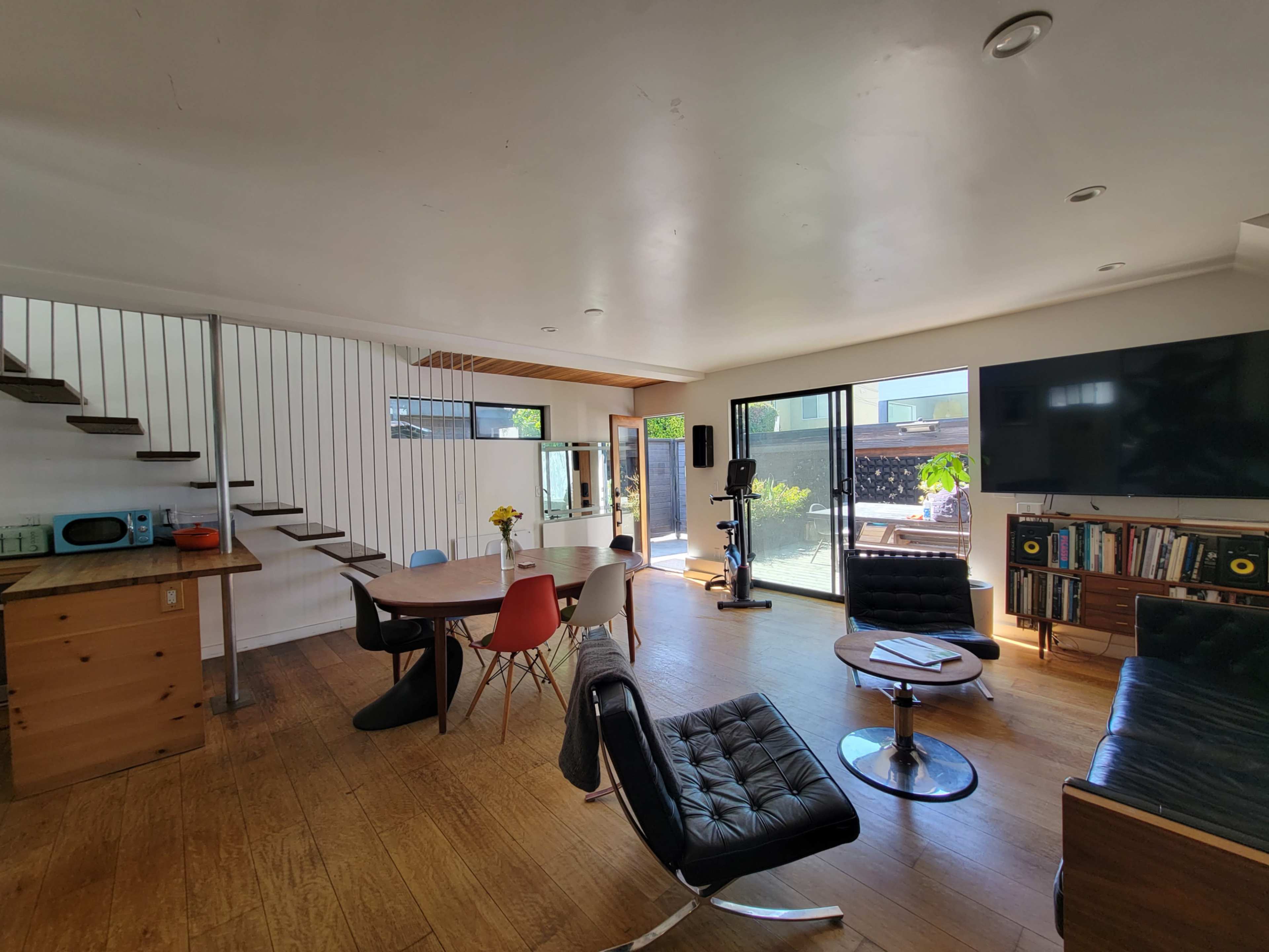 The image shows a modern living space featuring a dining area with a round table, a kitchen section, a lounge area with black seating, and large windows opening to an outdoor patio.