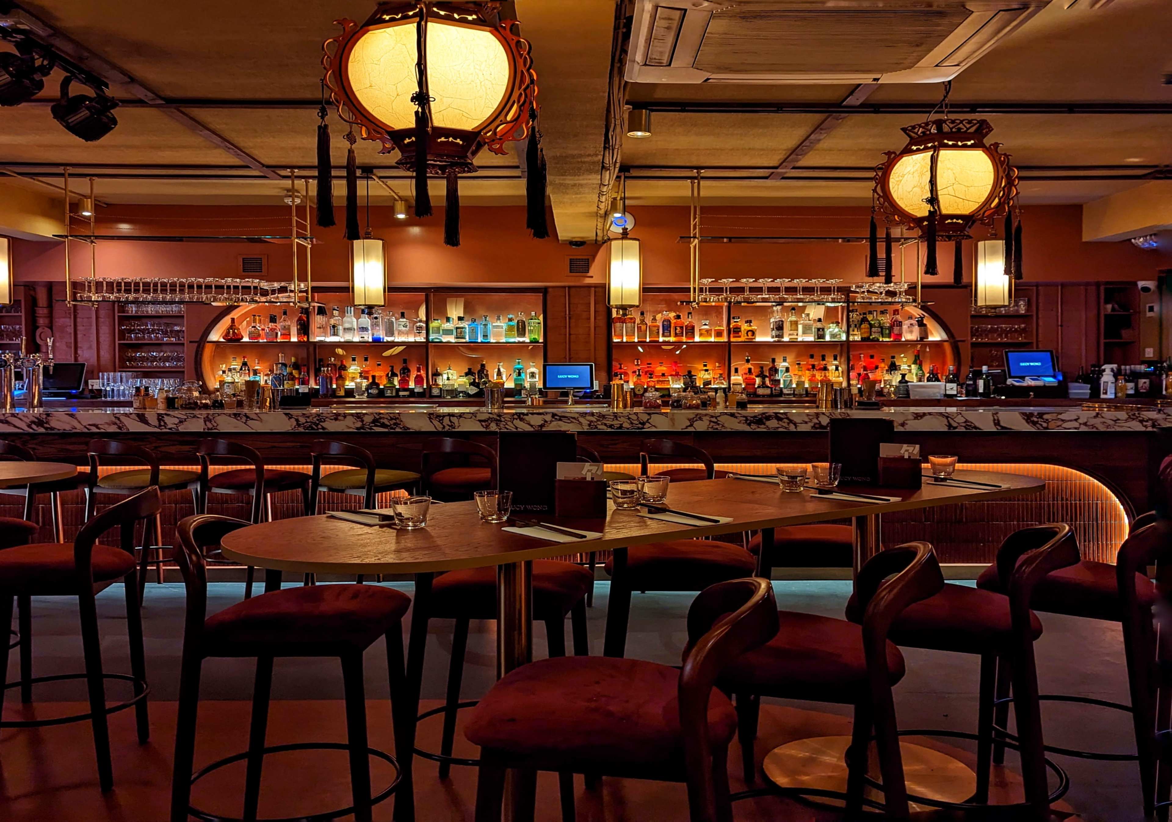 The image shows a stylish bar area with wooden tables and high stools, illuminated by decorative lanterns and featuring a well-stocked back bar.