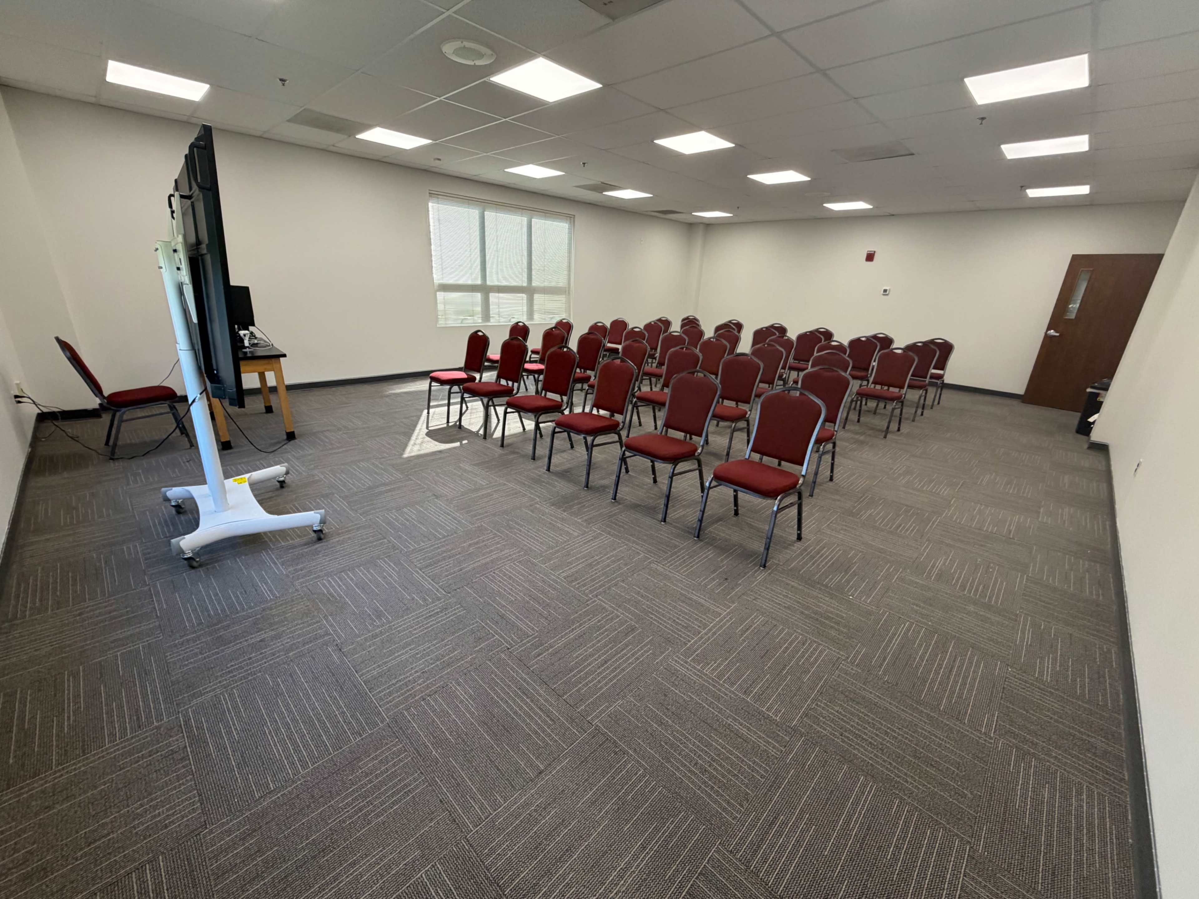 The image shows a sparsely furnished conference room with rows of red chairs facing a large display screen.