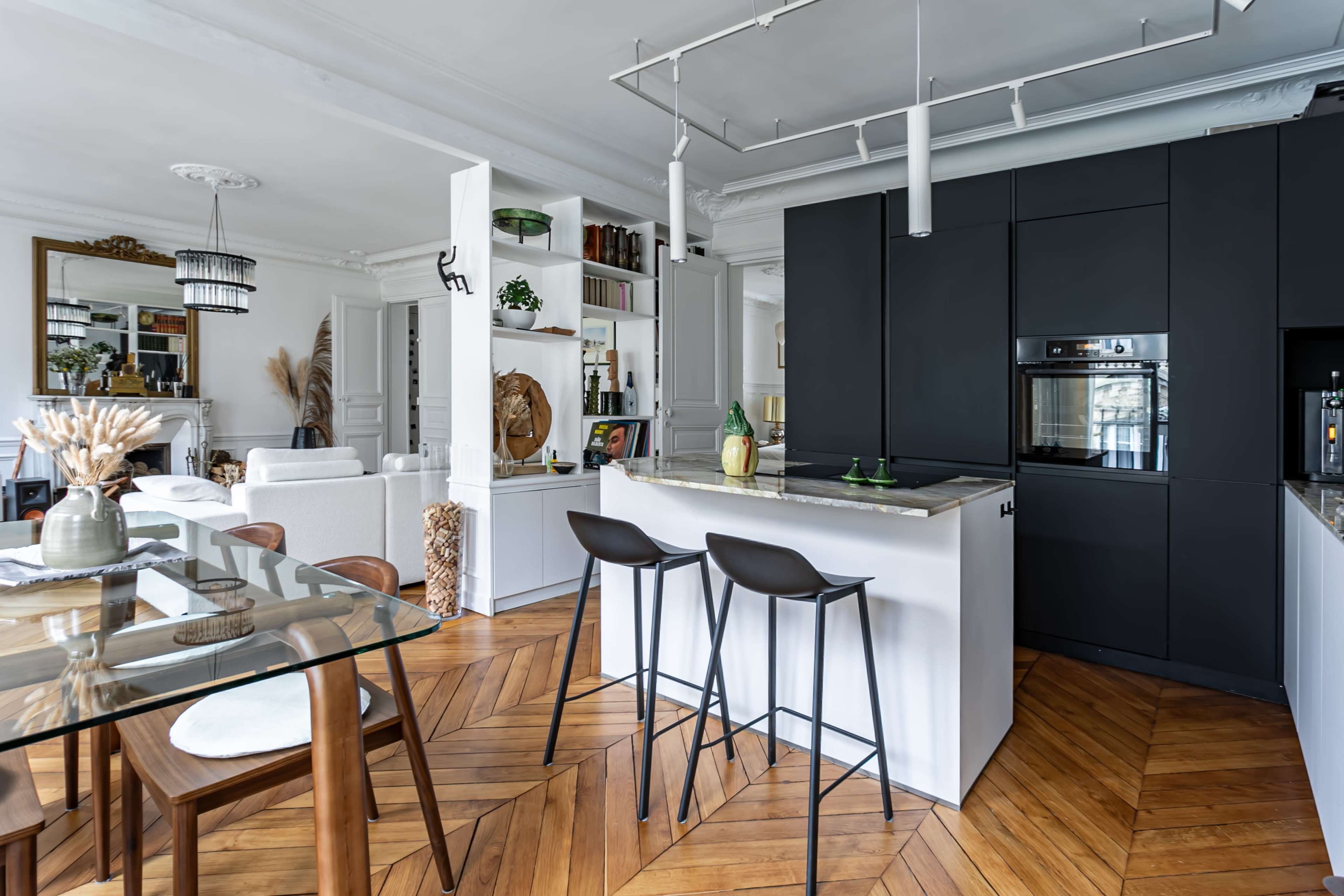A modern kitchen features a central island with bar stools, surrounded by wooden flooring, a dining table, and stylish cabinetry.