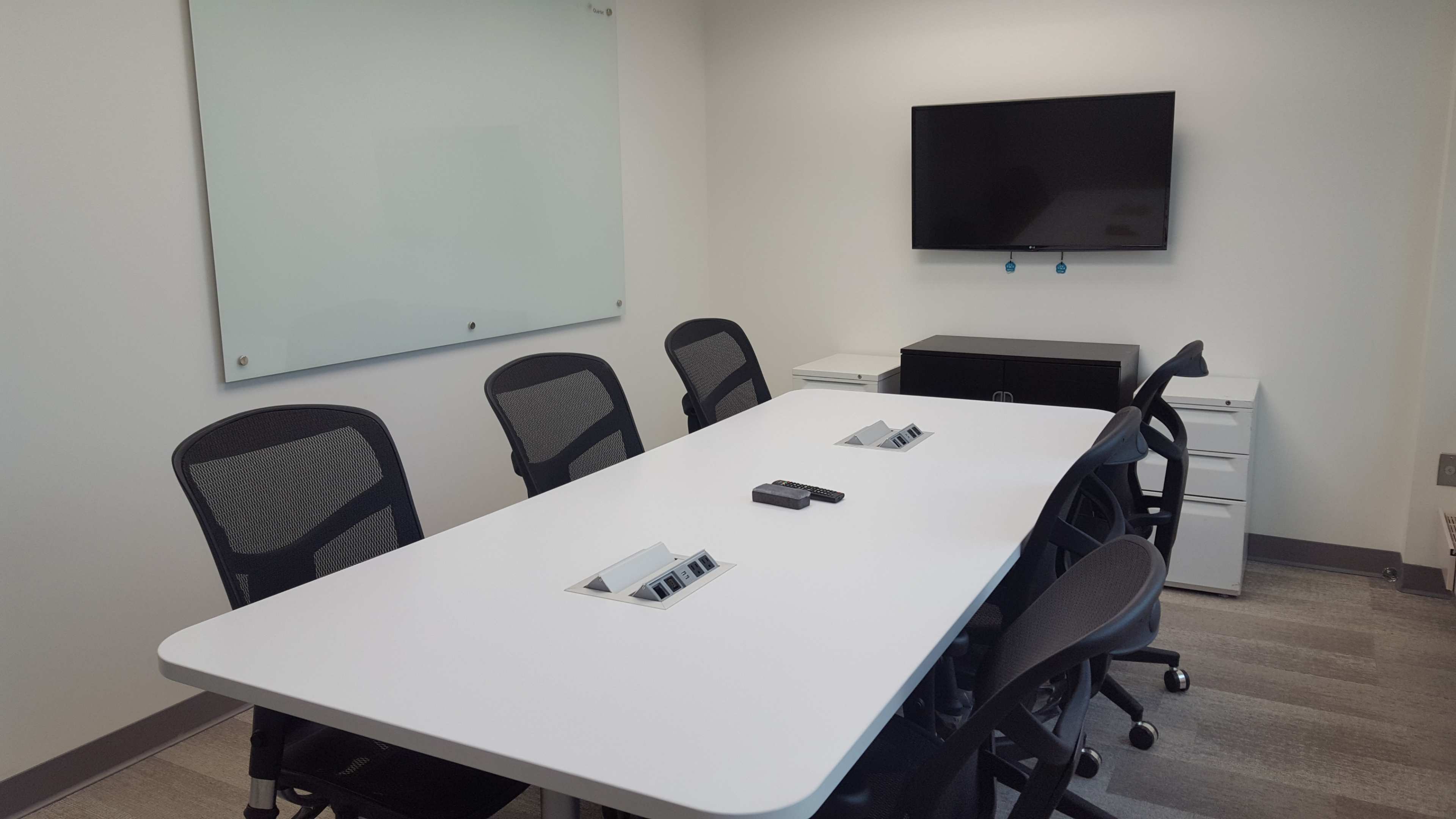 A conference room features a long white table with several black chairs, a wall-mounted television, and a whiteboard.