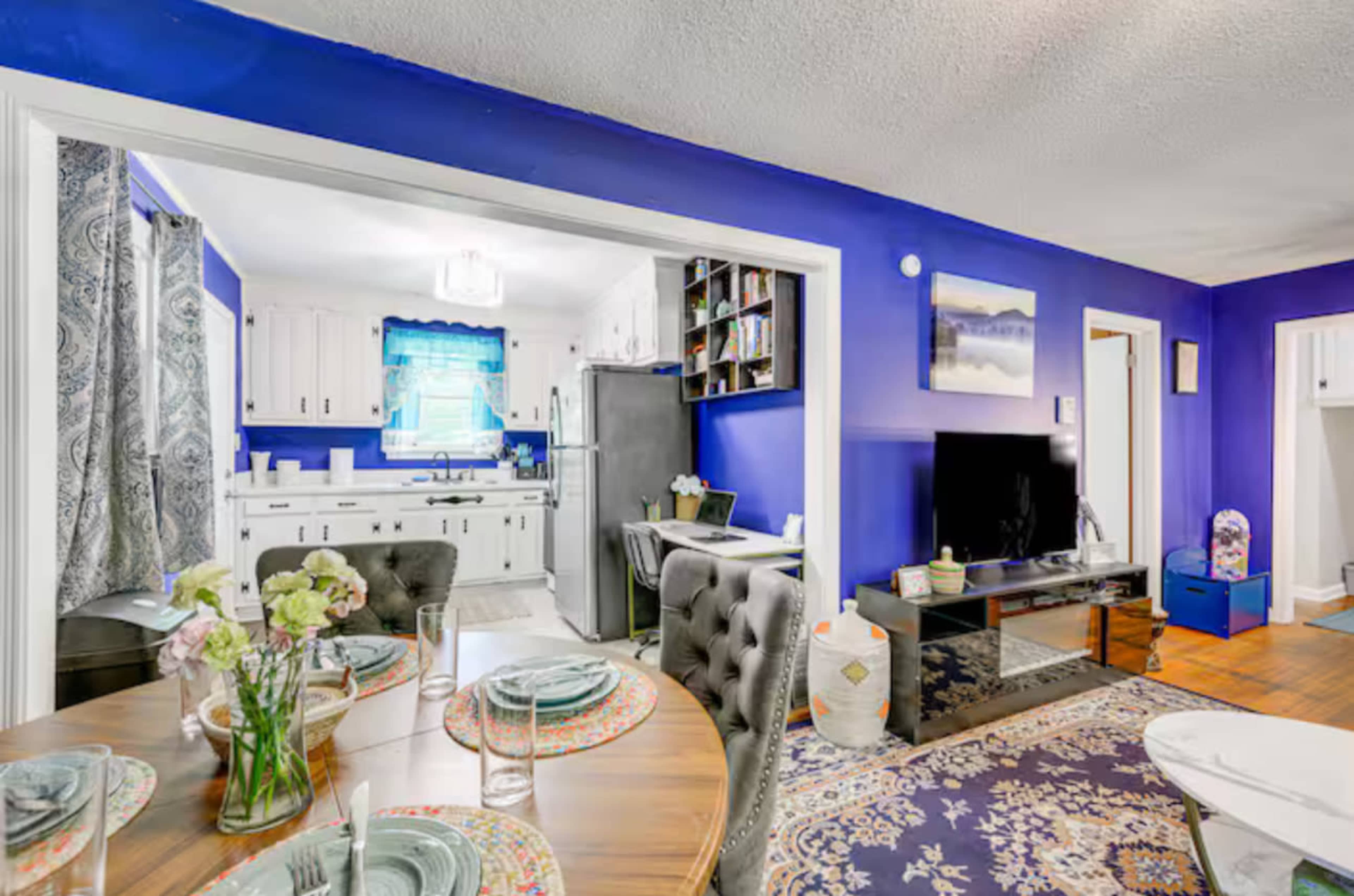 The image shows a dining area with a round table set for a meal, adjacent to a kitchen with white cabinets and a blue accent wall.