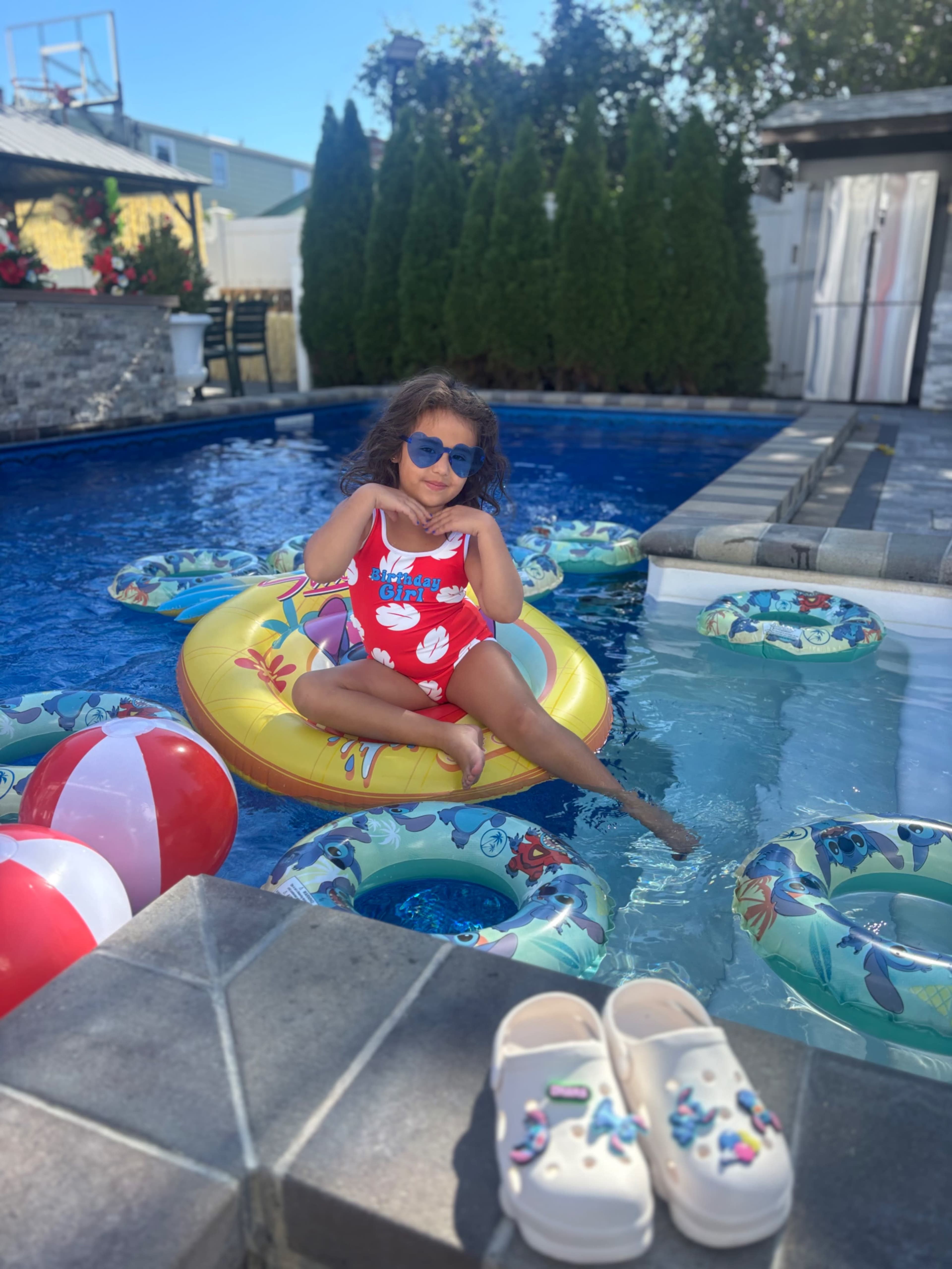 A child in a red swimsuit relaxes on a yellow inflatable float in a pool surrounded by additional colorful floats and beach balls.