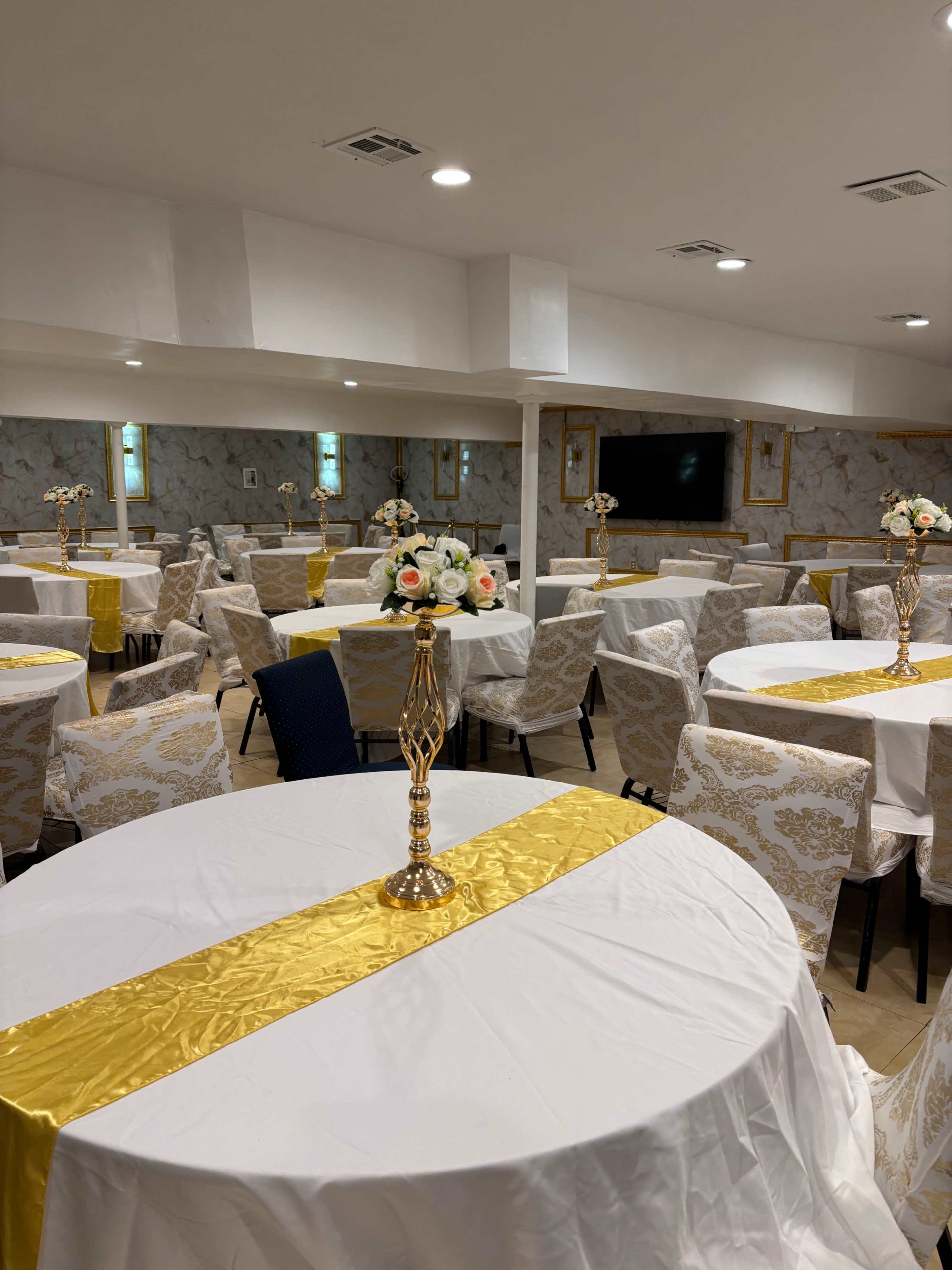The image shows a banquet hall set with white tables covered by tablecloths, each adorned with golden table runners and centerpieces of flowers.