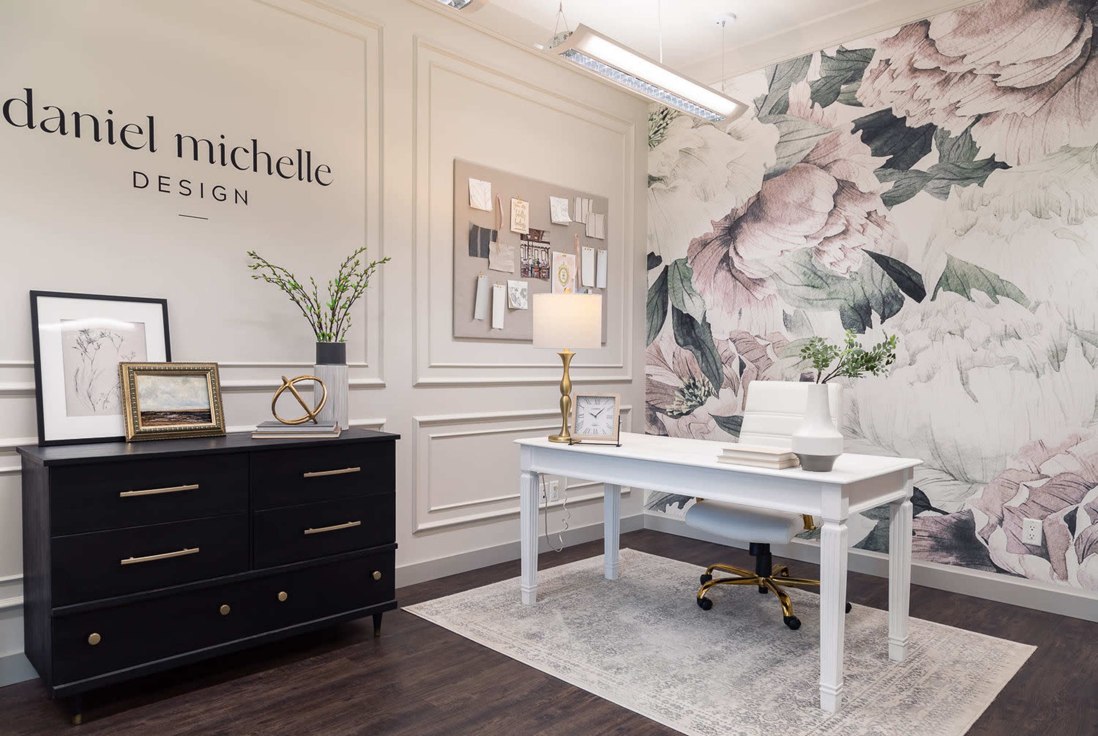 A modern office space featuring a white desk, a black dresser, floral wallpaper, and decor elements including a lamp and framed pictures.