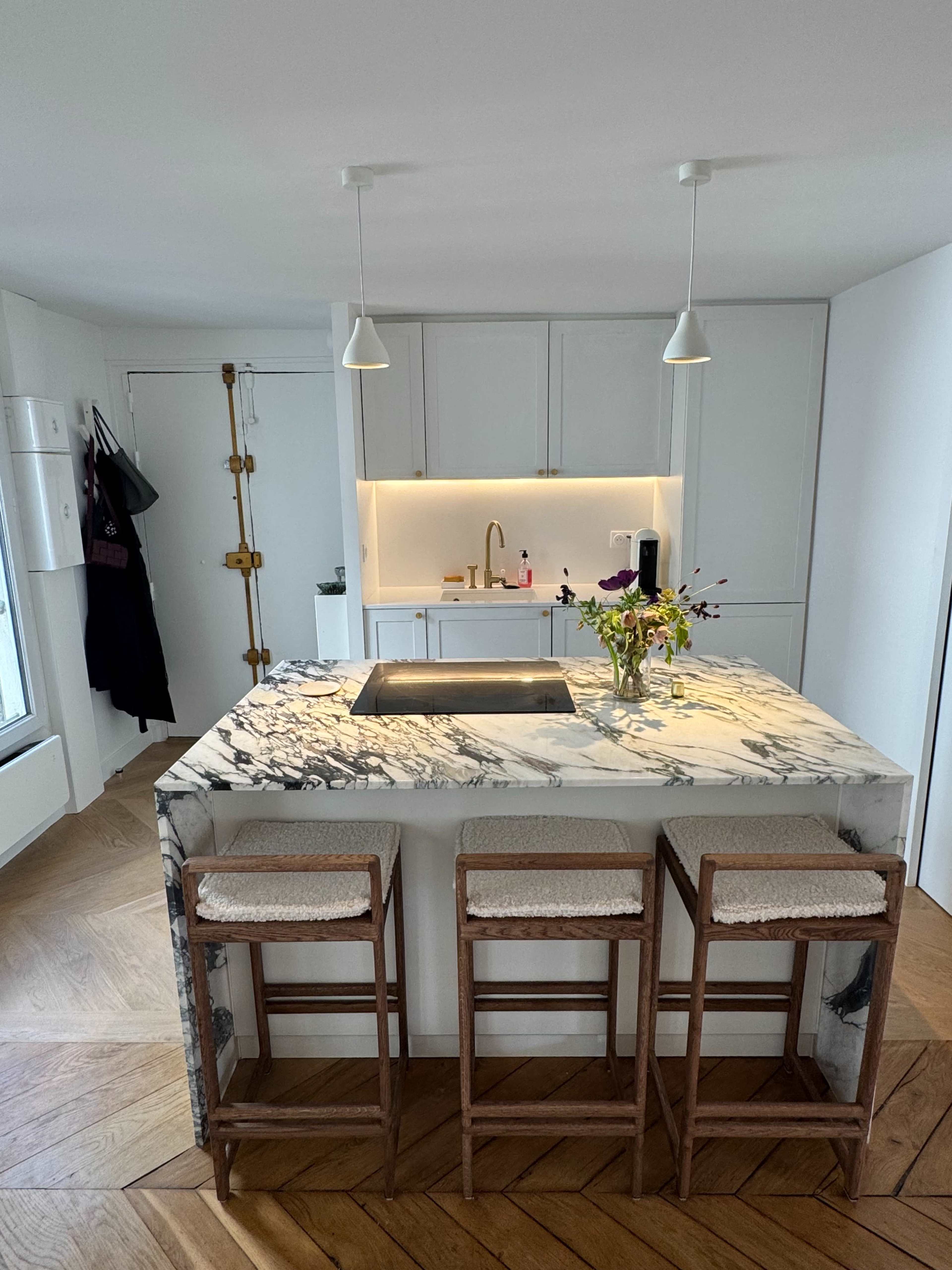 The image shows a modern kitchen with a large marble island and three wooden bar stools.