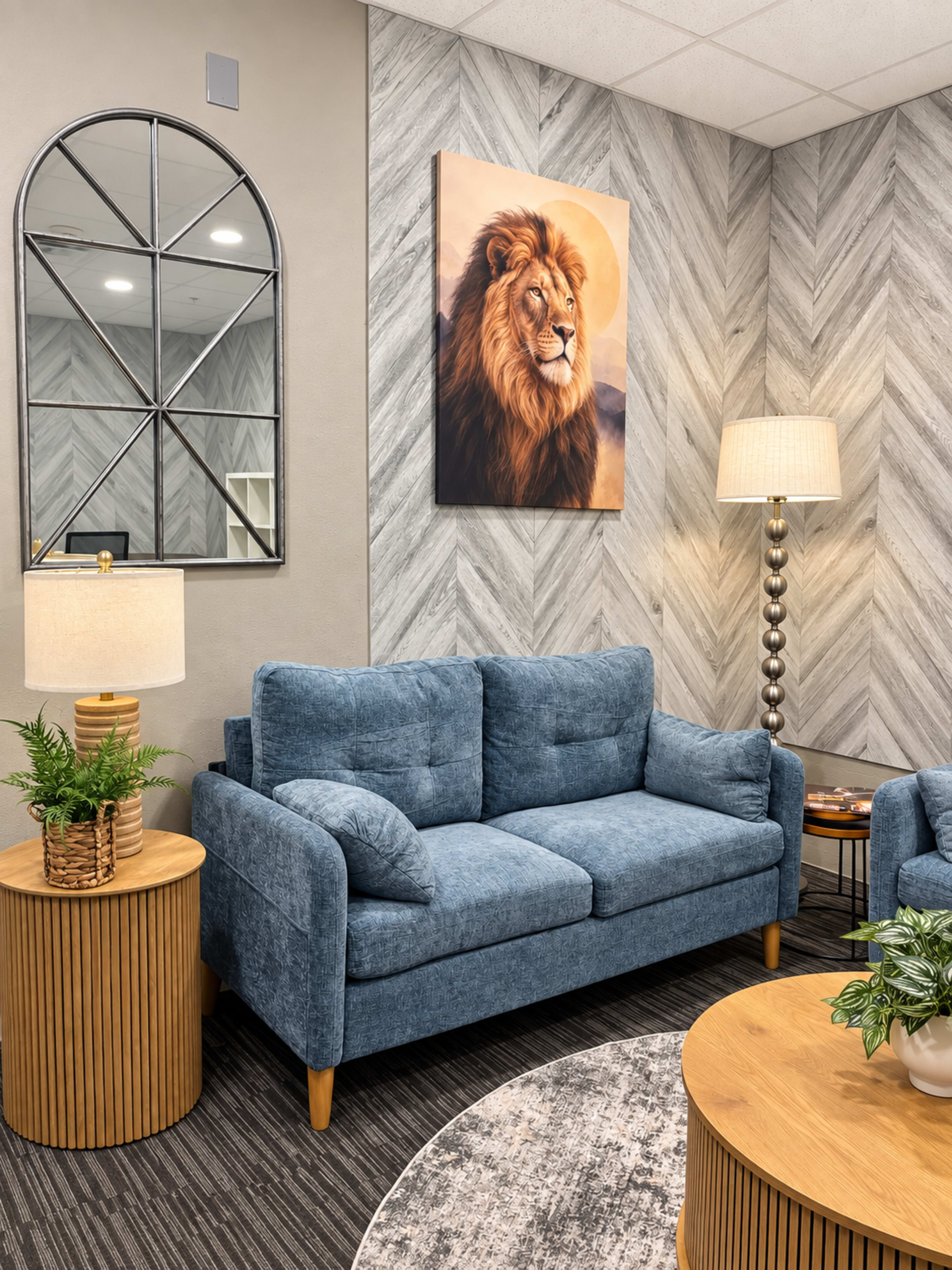 The image shows a cozy sitting area featuring a blue couch, a round wooden table, a lamp, and a wall with a lion painting and a large mirror.