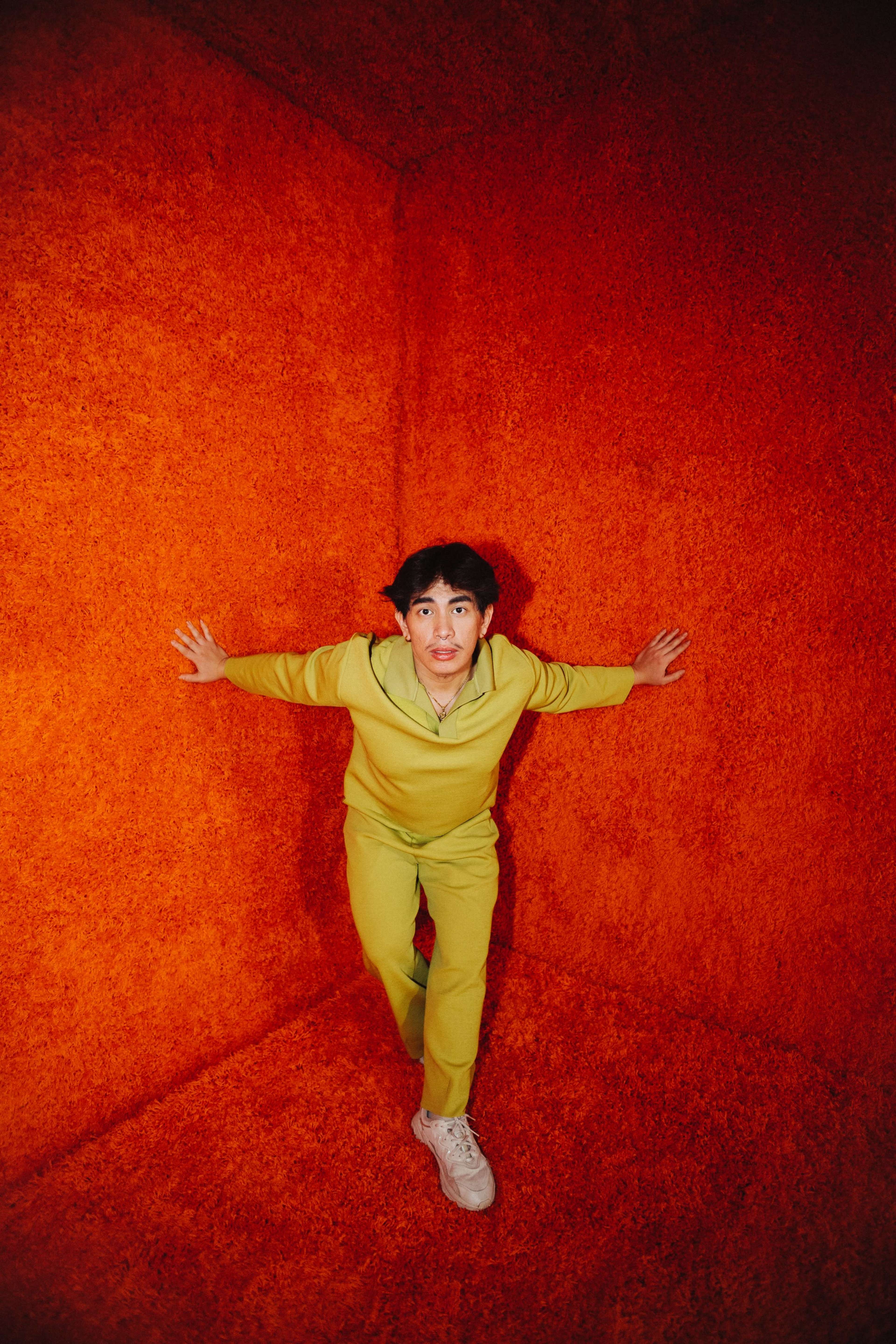 A person in a yellow tracksuit leans against an orange-textured wall inside a small, enclosed space.