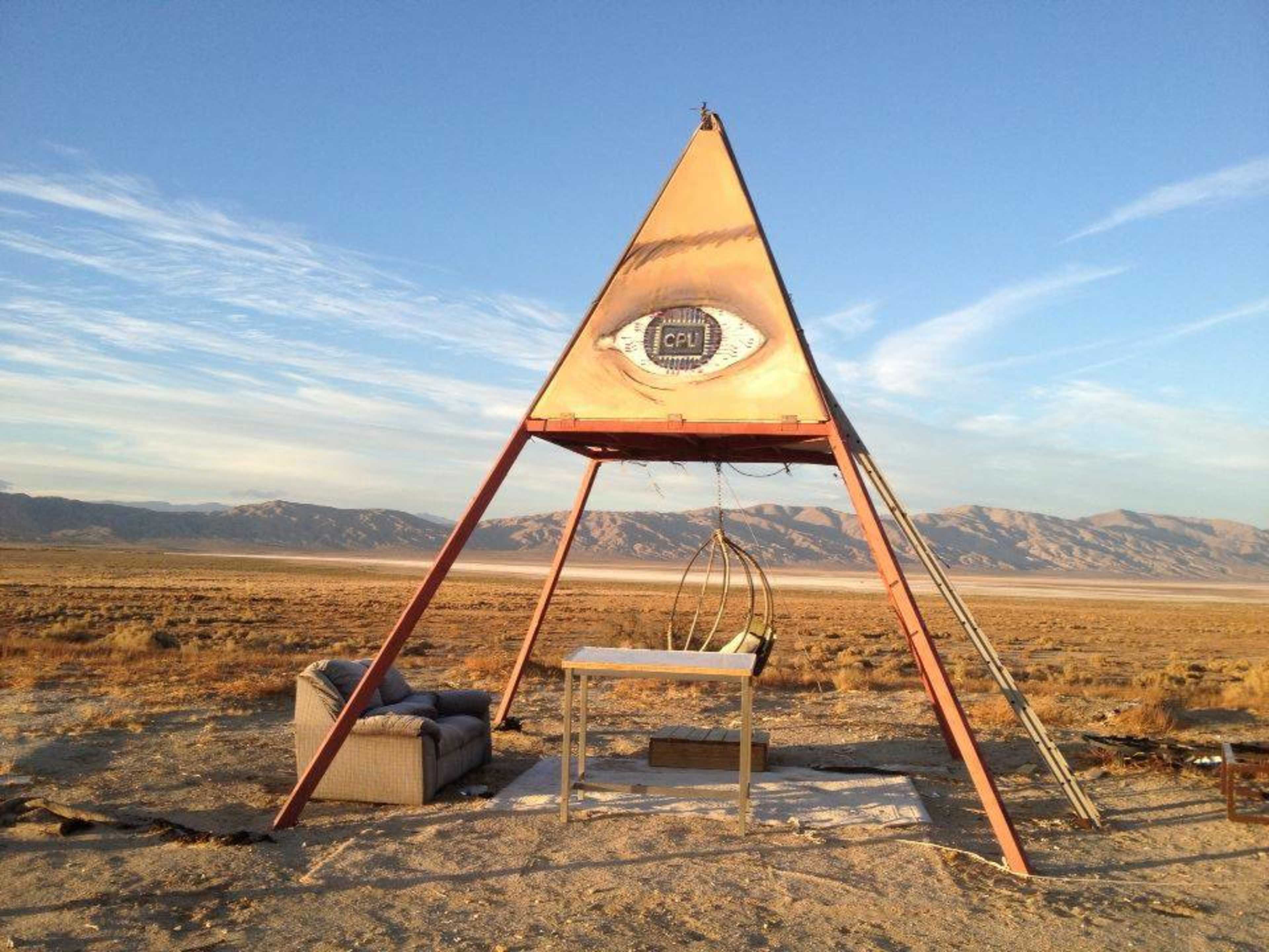 An unconventional structure resembling a pyramid features a hanging seat and a table in a barren landscape with mountains in the background.