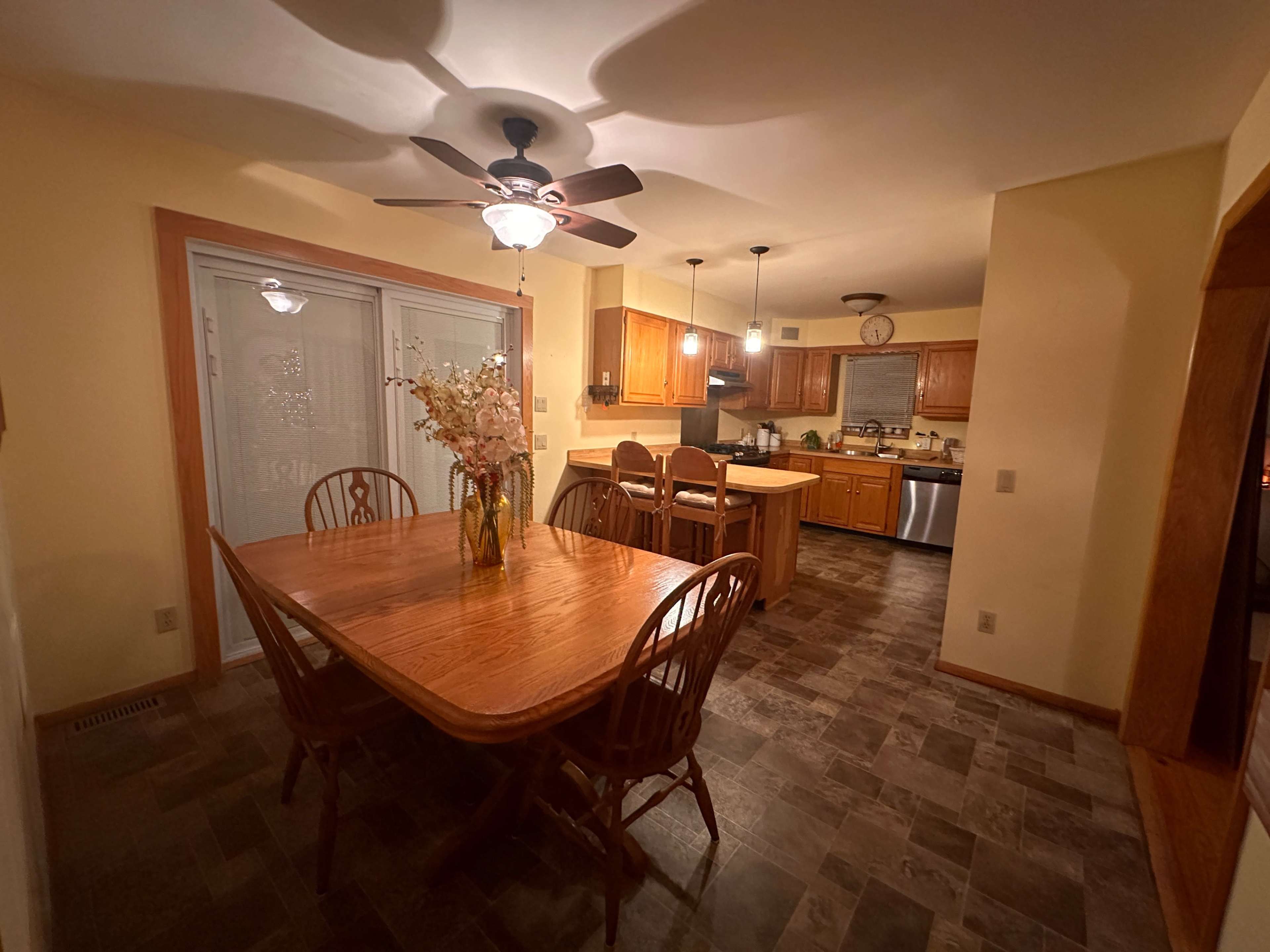 The image shows a dining area with a wooden table and chairs, adjacent to a kitchen with wooden cabinetry and modern appliances.