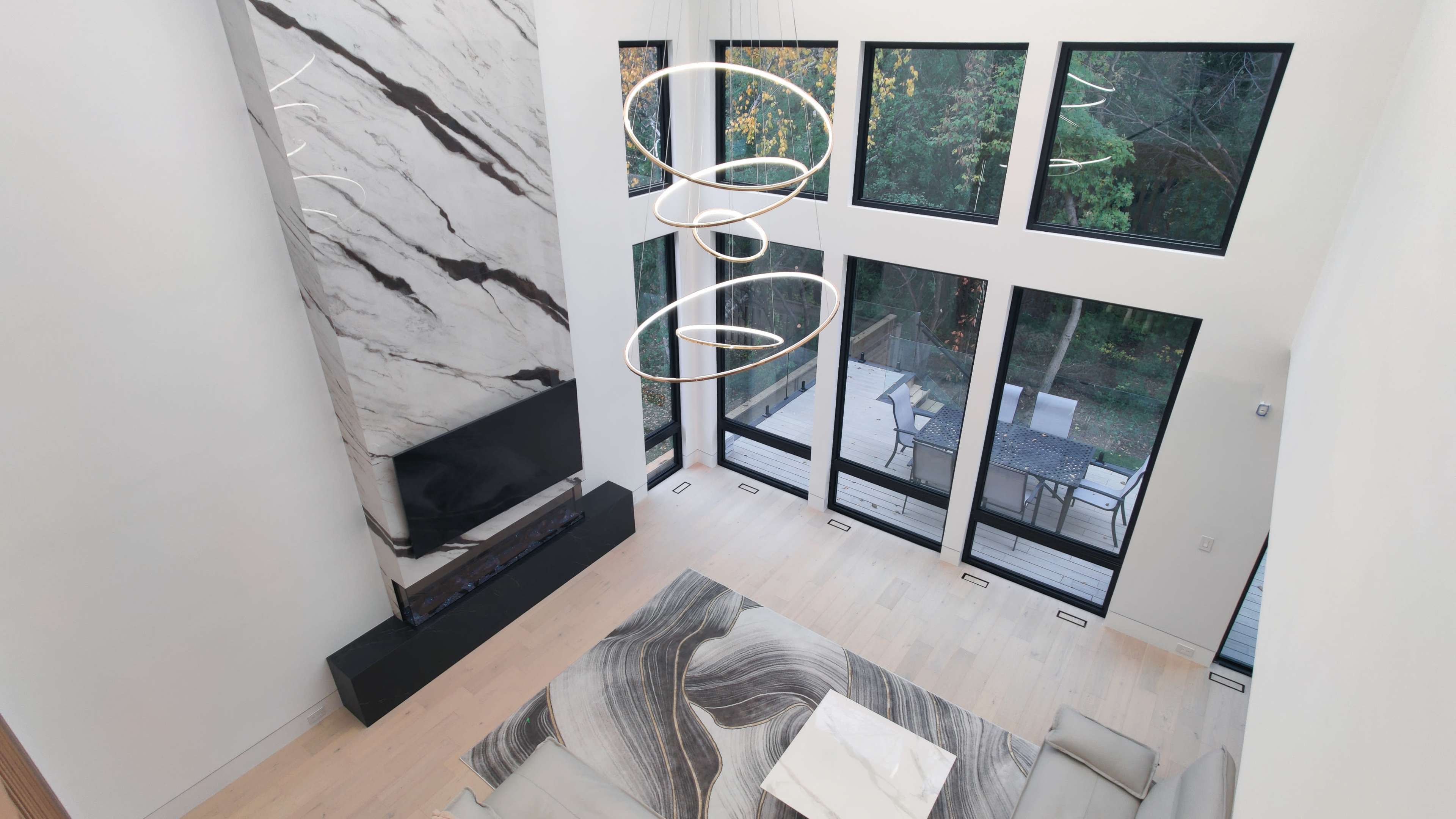 A modern living room with a marble accent wall, large windows overlooking a wooded area, and contemporary furniture arranged around a patterned rug.