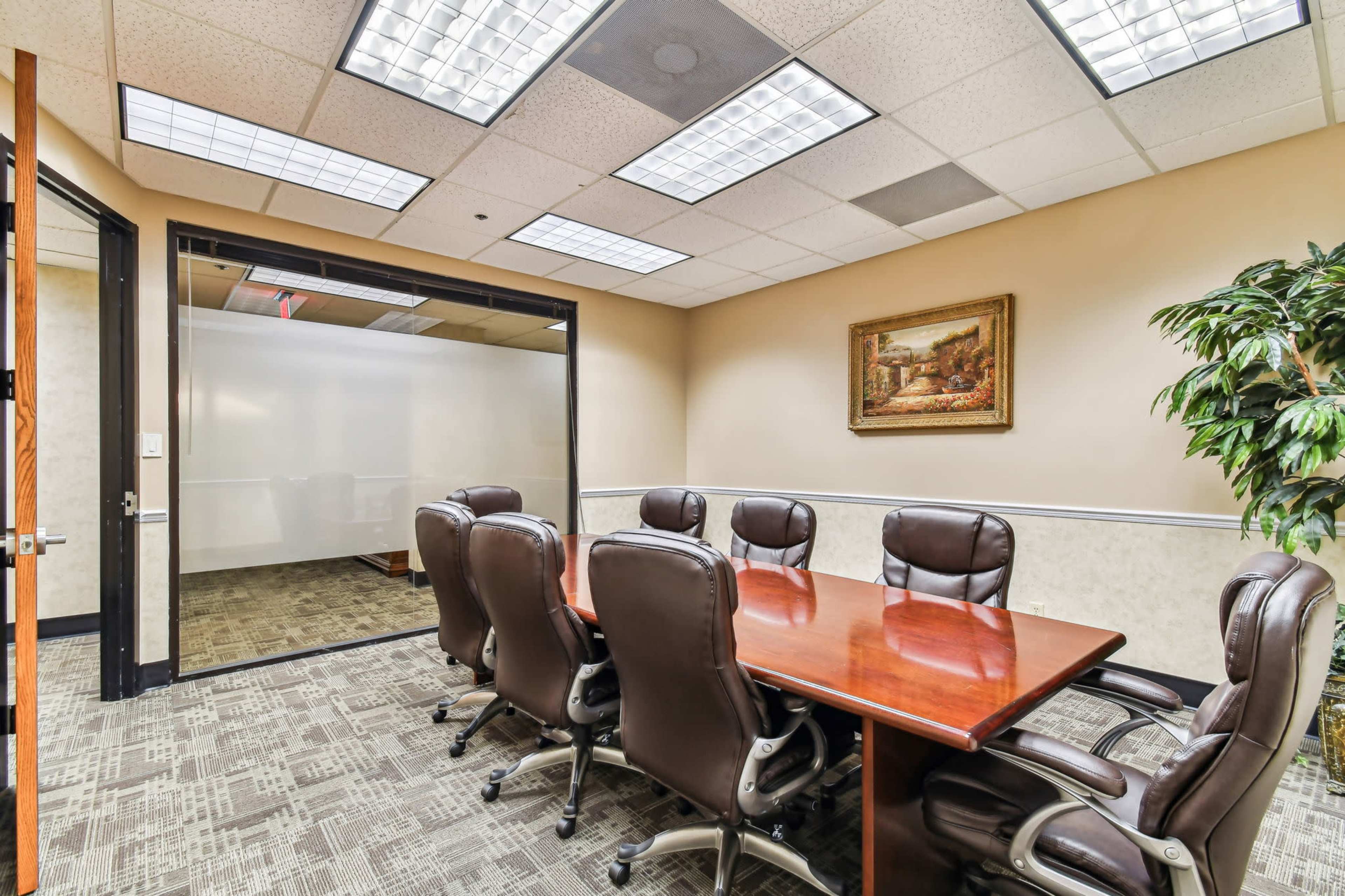 The image shows a conference room with a large wooden table surrounded by leather chairs, and a glass-walled meeting room in the background.