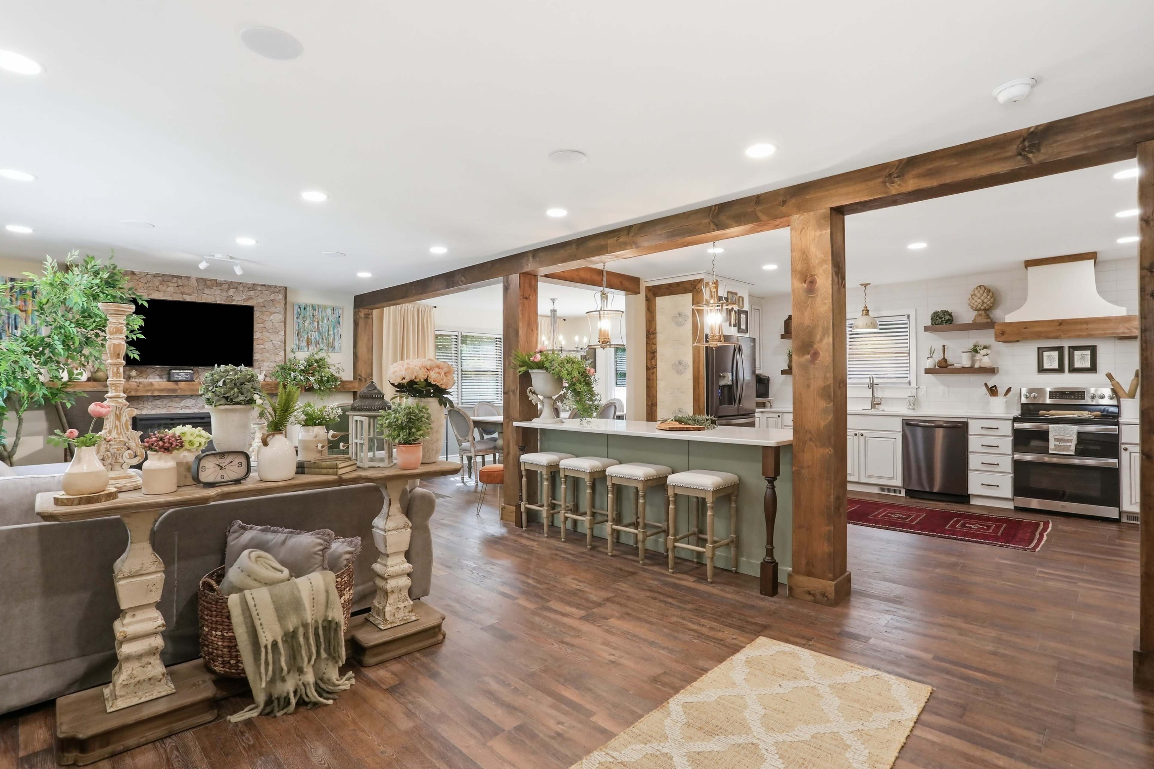 The image shows a modern, open-concept living space that includes a kitchen and a seating area, with wooden beams, plants, and decorative accessories.