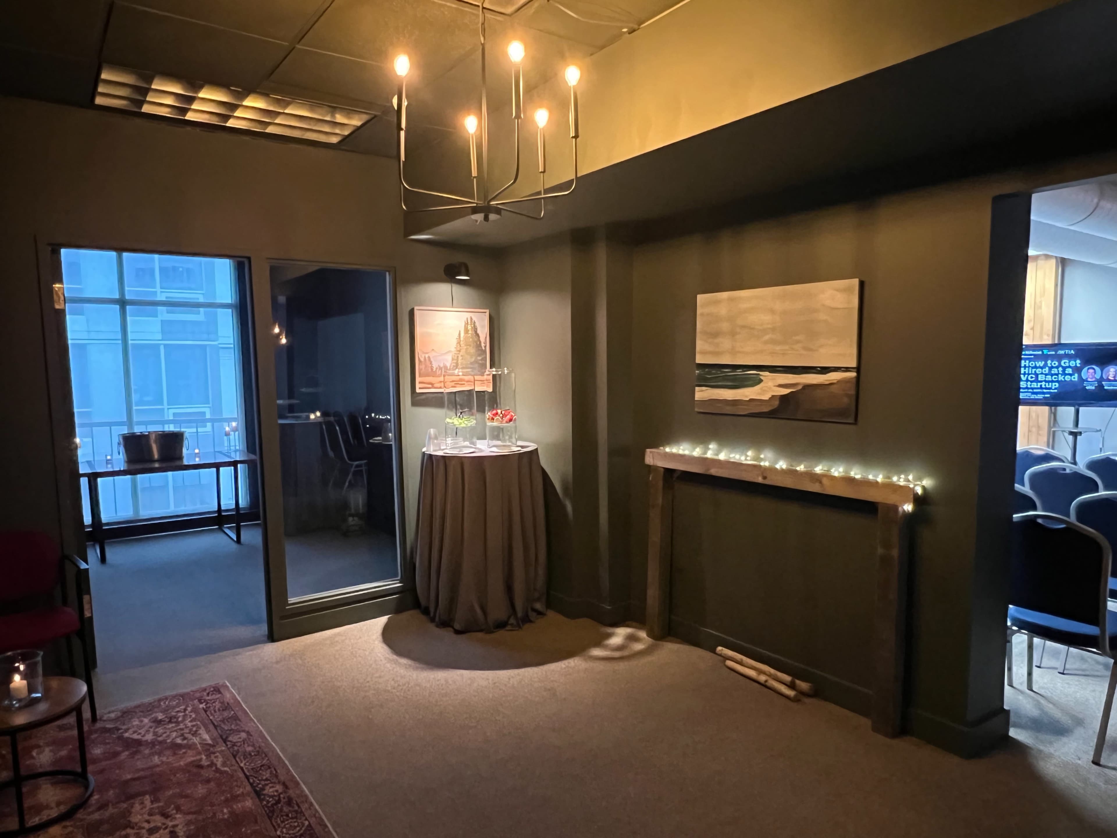 A dimly lit room with a small dining table covered by a gray cloth, a modern chandelier, and an artwork depicting waves on the wall.