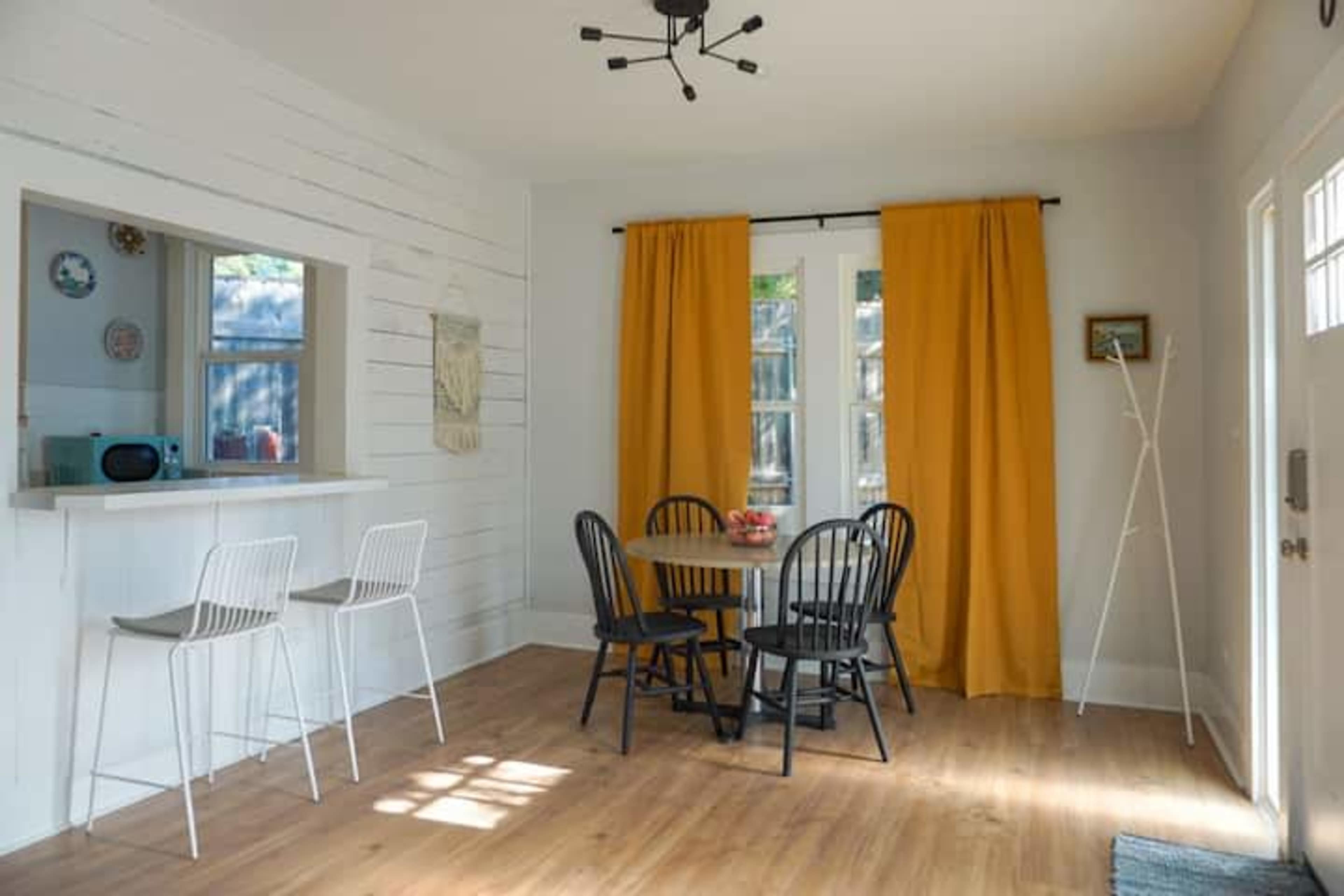 A bright, minimalist dining area features a circular black table surrounded by black chairs, with yellow curtains framing a window and a small bar counter in the background.