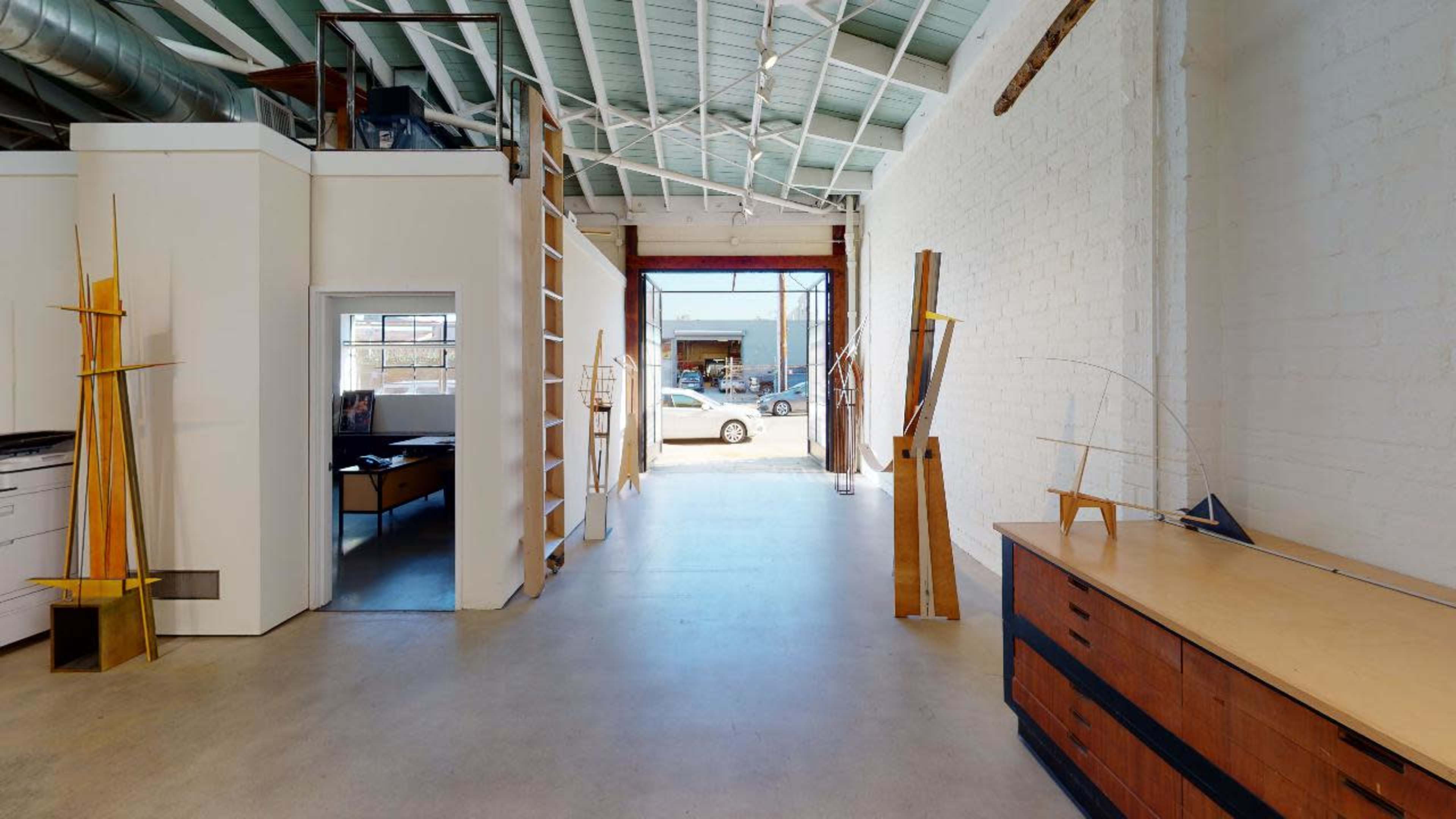 A spacious, well-lit studio with a wooden desk, shelves, and artistic sculptures, leading to an open entryway that overlooks a car parked outside.