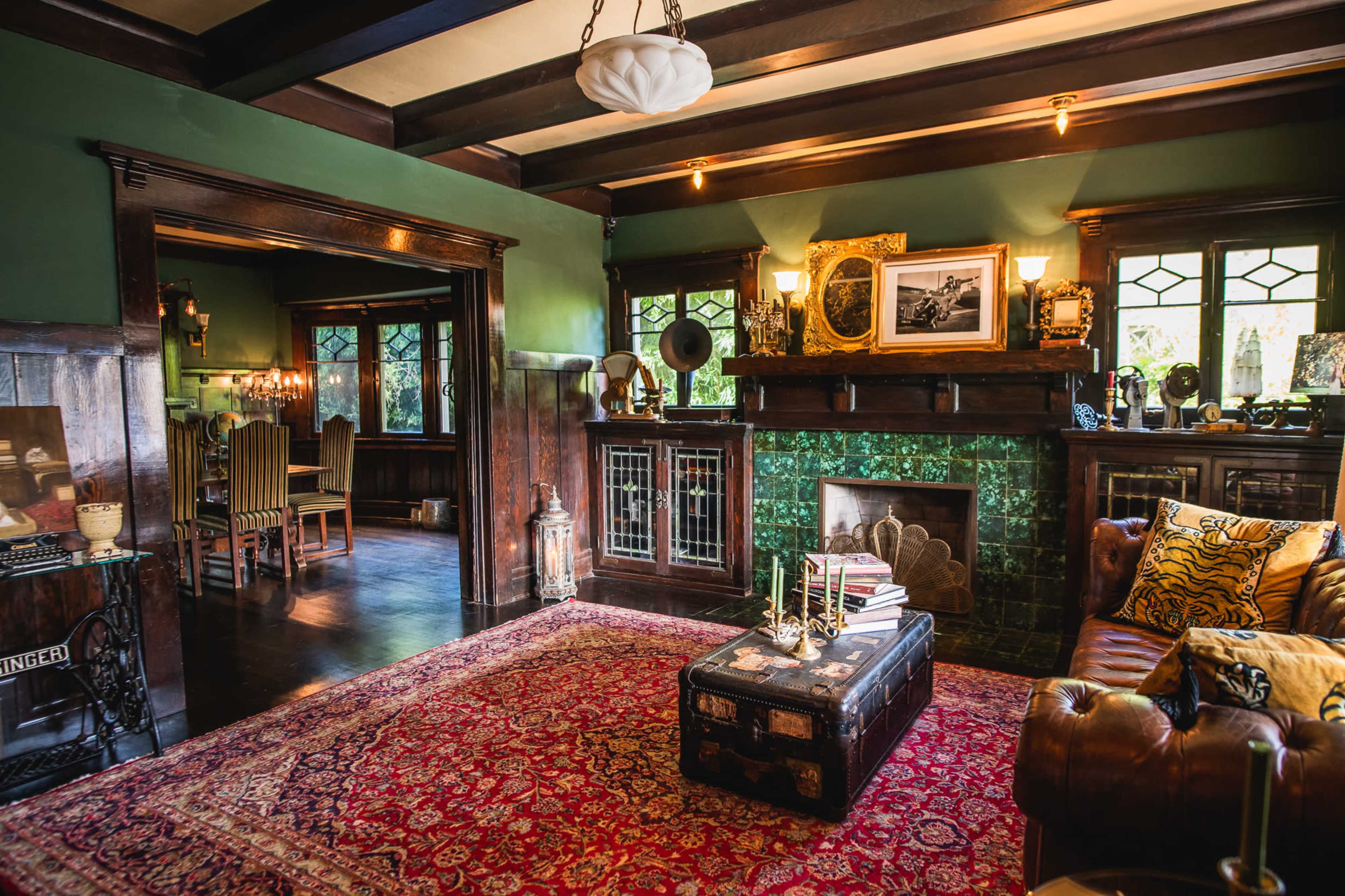 A cozy living room featuring a wooden fireplace with green tiles, a decorative red area rug, a coffee table, and vintage decor elements.