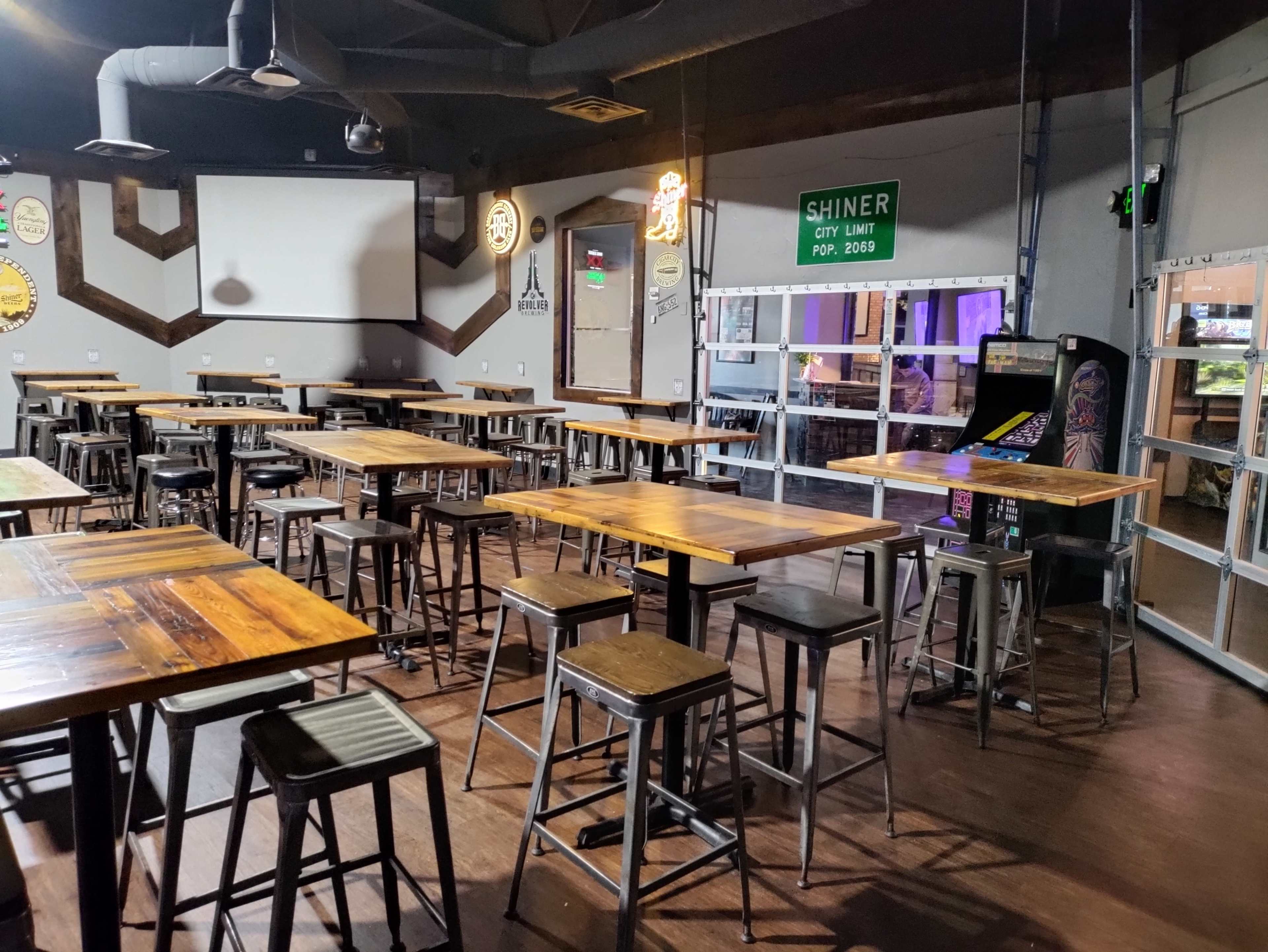 An empty dining area with wooden tables and metal stools, featuring a bar arcade machine in the corner and a large screen on the wall.