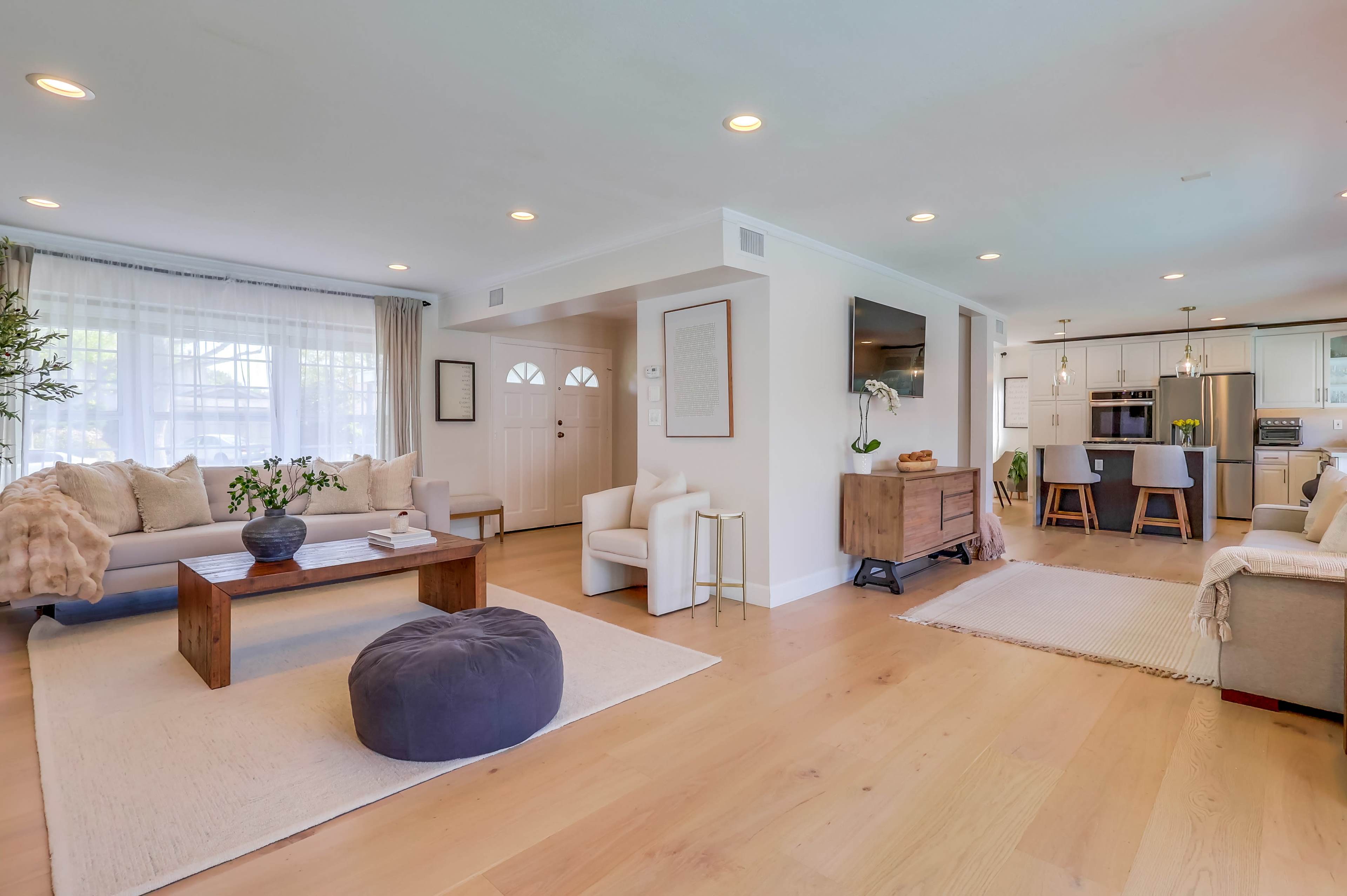 The image shows a modern living room with a light color scheme, featuring a sofa, a coffee table, a pouf, and a dining area connected to a kitchen.