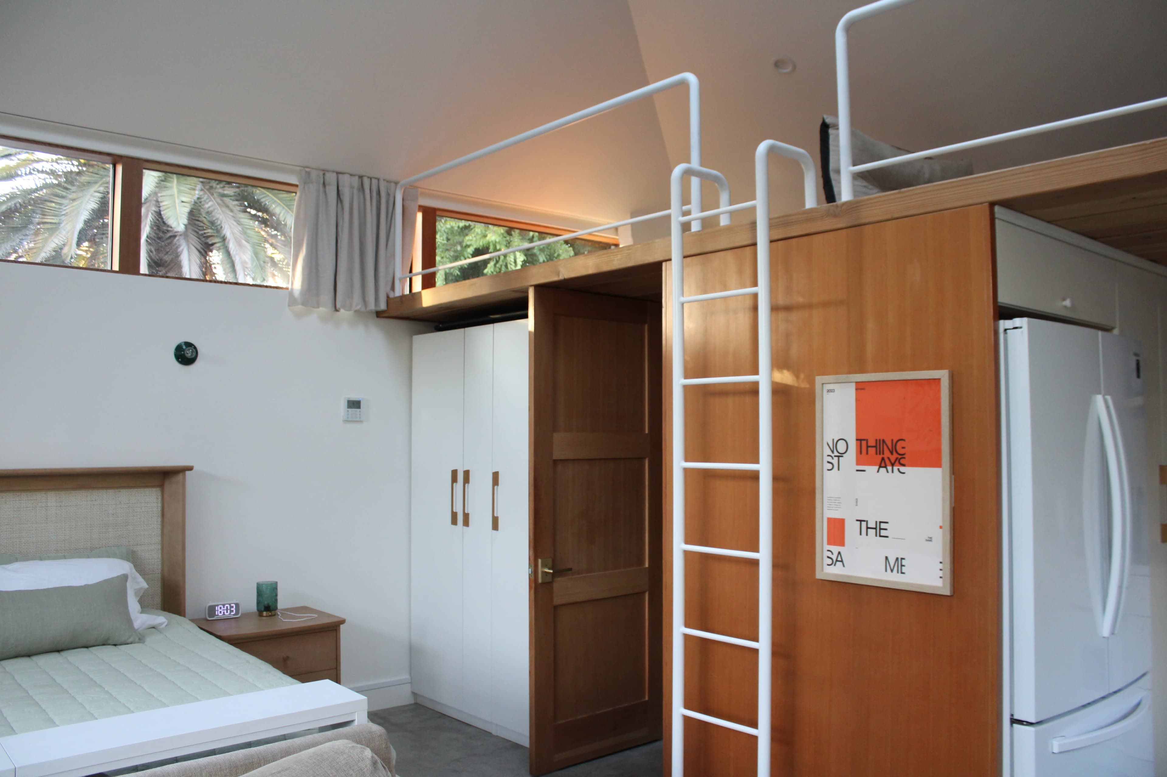 A modern bedroom features a lofted sleeping area accessible by a ladder, with a bed, wardrobe, and large windows letting in natural light.
