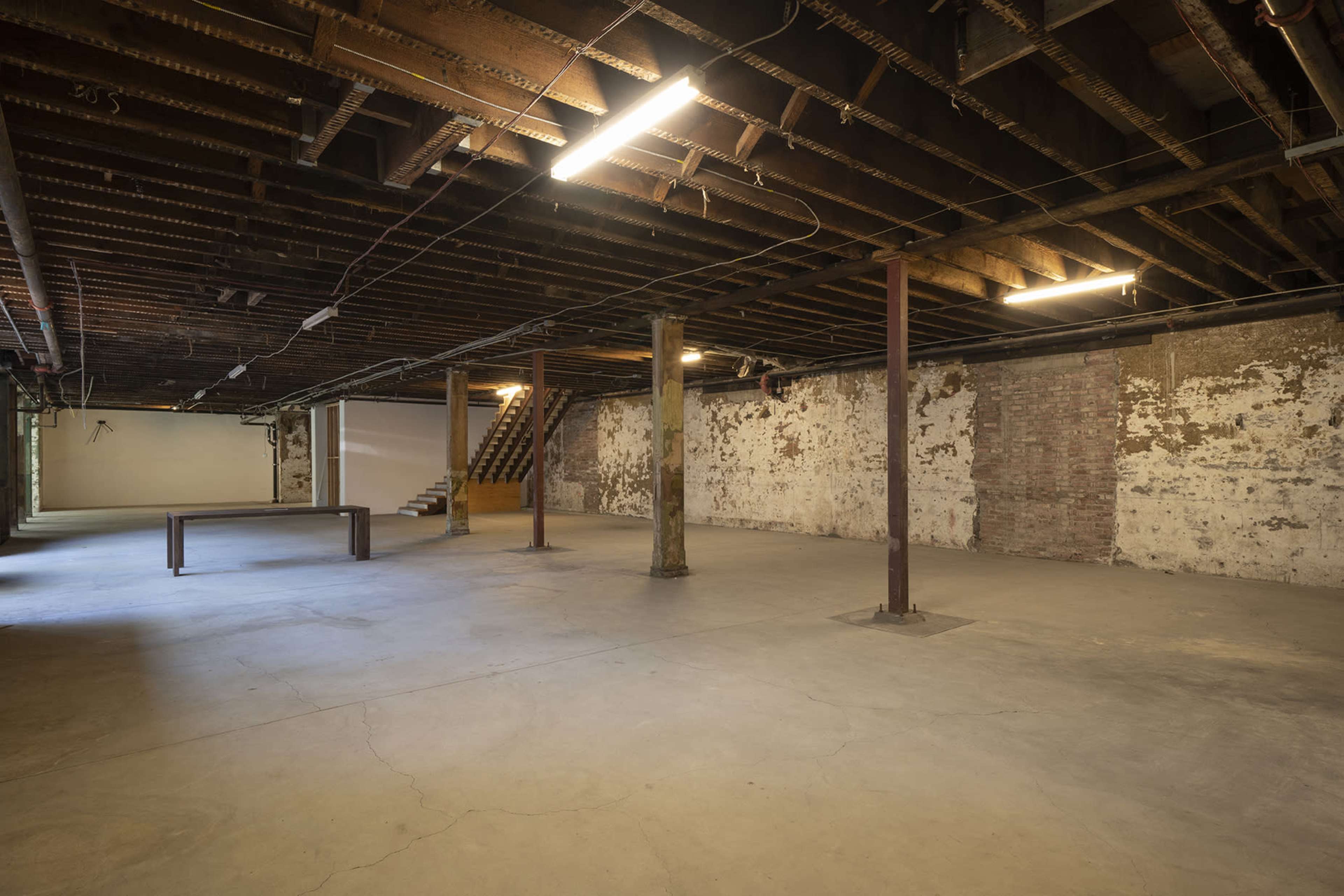 A large, empty basement with exposed beams, concrete flooring, and a staircase leading to an upper level.