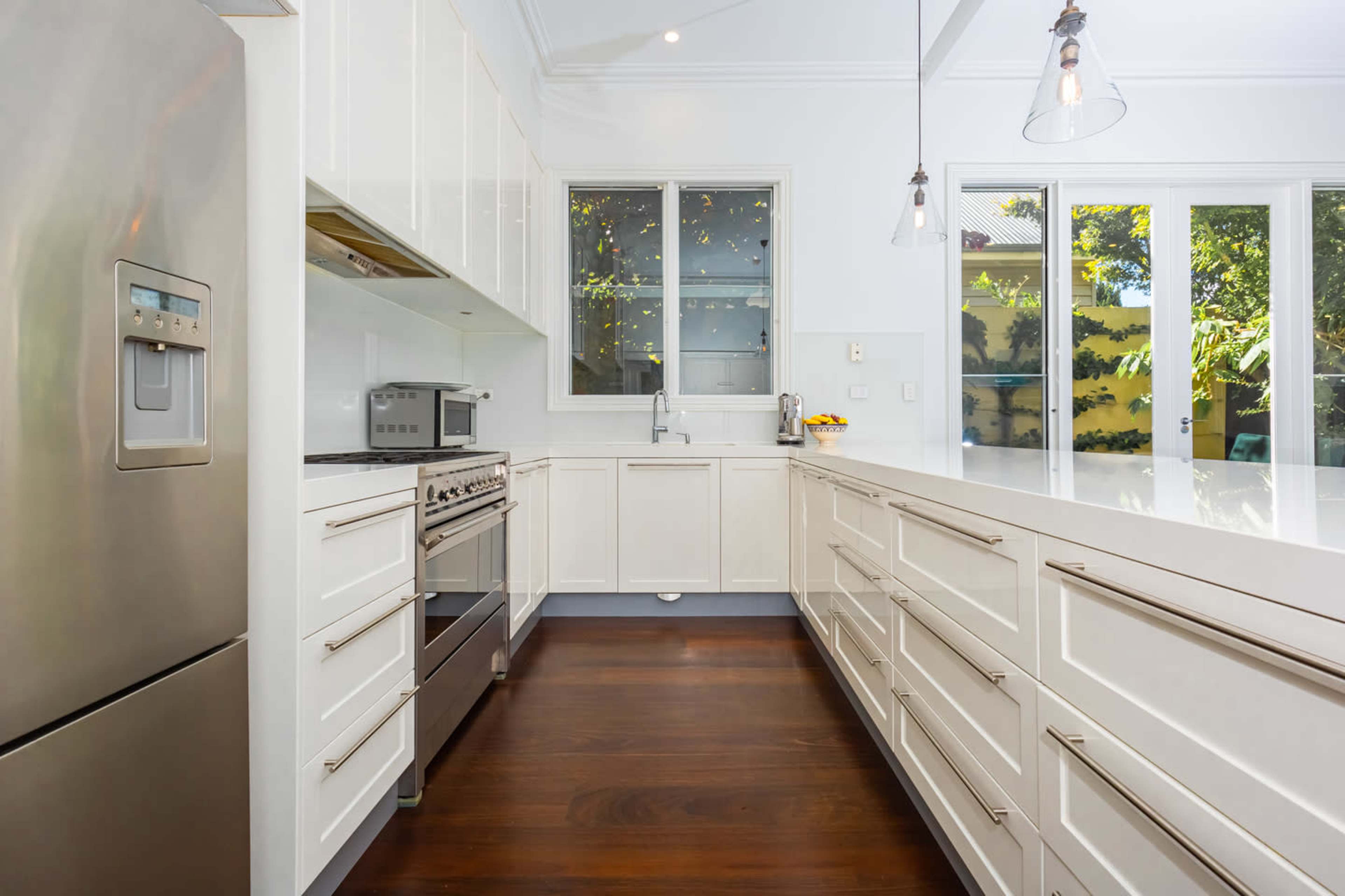 The image shows a modern kitchen featuring white cabinets, stainless steel appliances, and a spacious countertop with large windows allowing natural light.