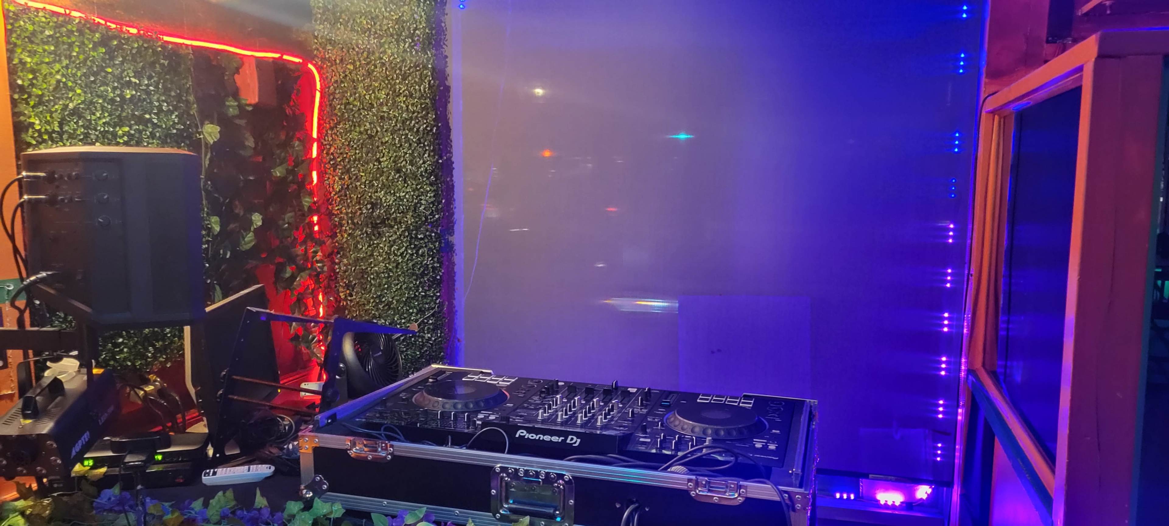 The image shows a DJ setup with a Pioneer mixer and equipment in a neon-lit space surrounded by greenery.